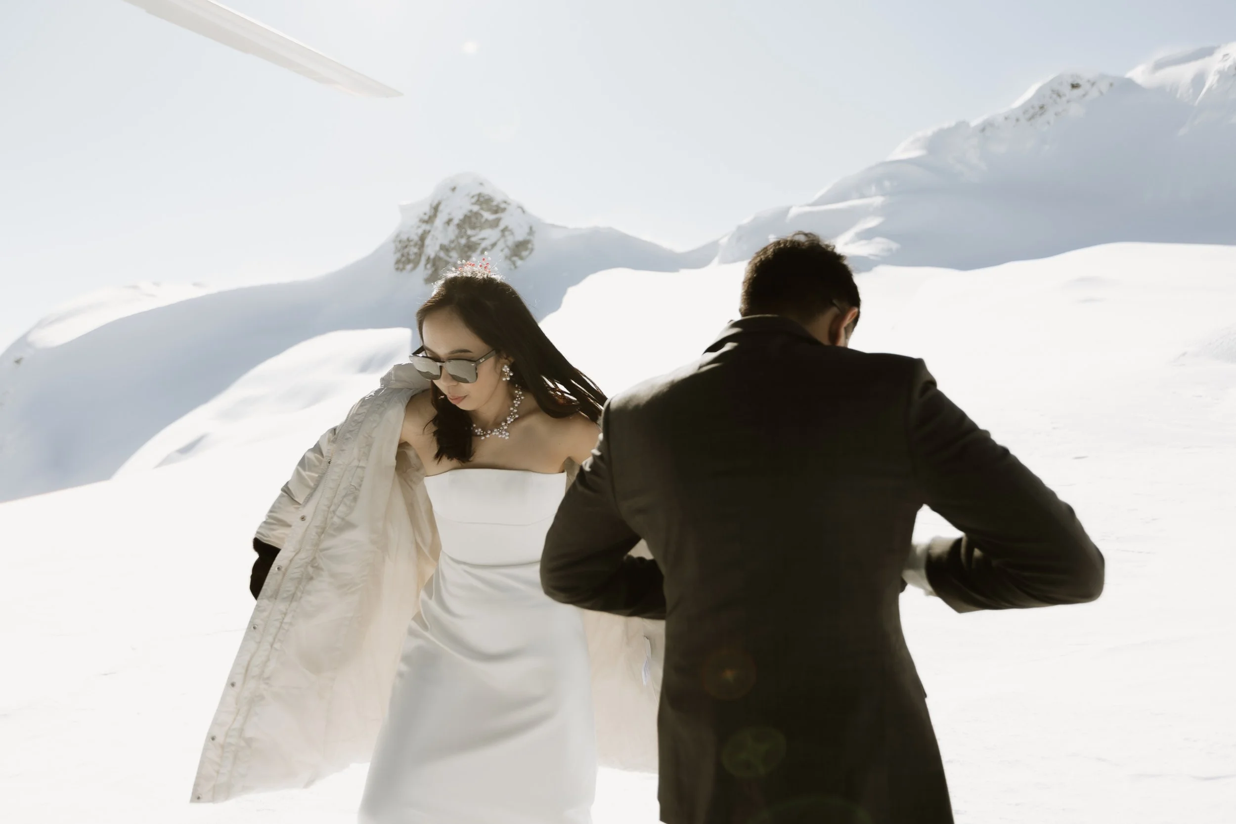 A woman in a white strapless dress and sunglasses, and a man in a black suit, standing in a snowy mountain landscape.