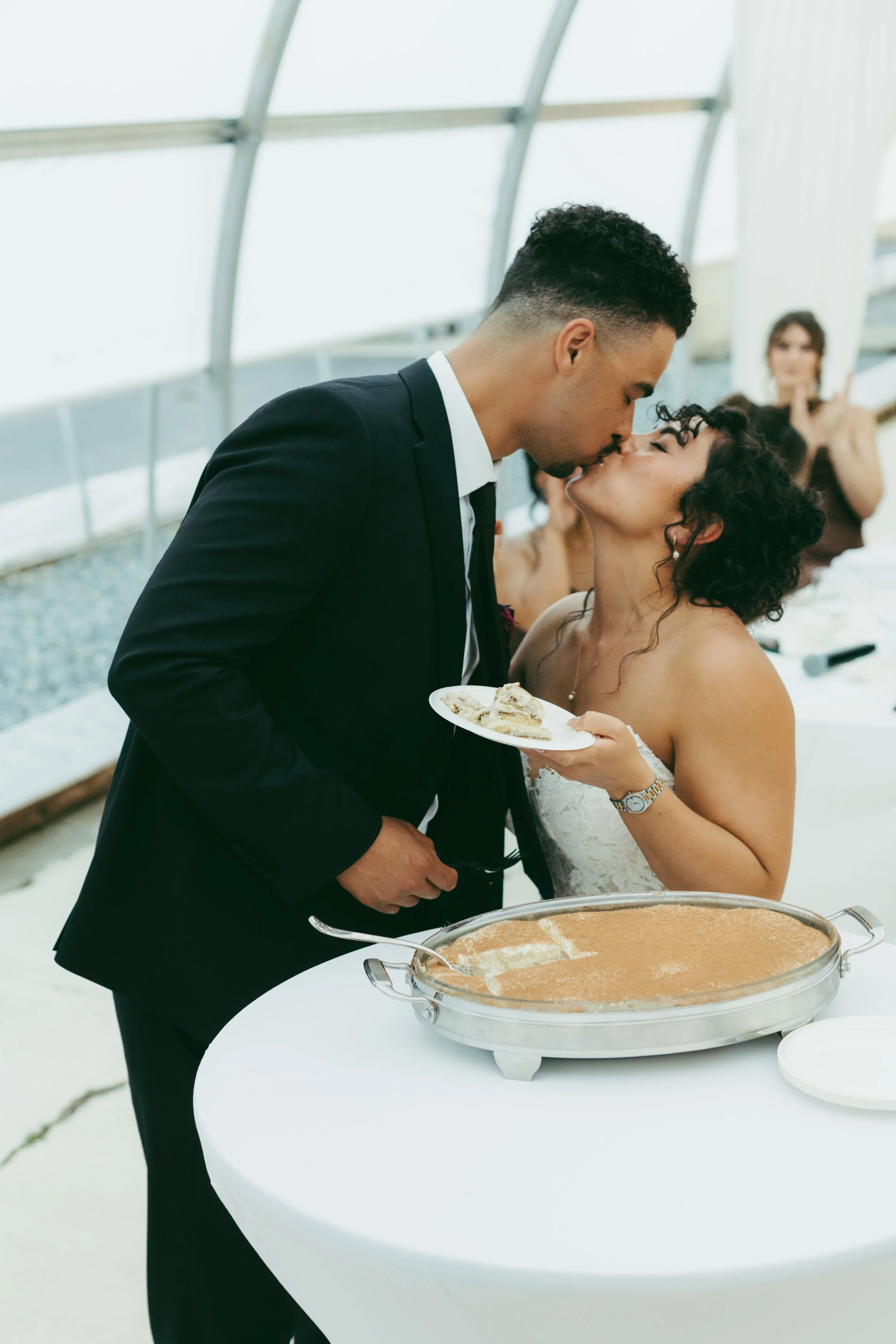 Bride and groom kiss beside a wedding dessert table with a large tray of cake.