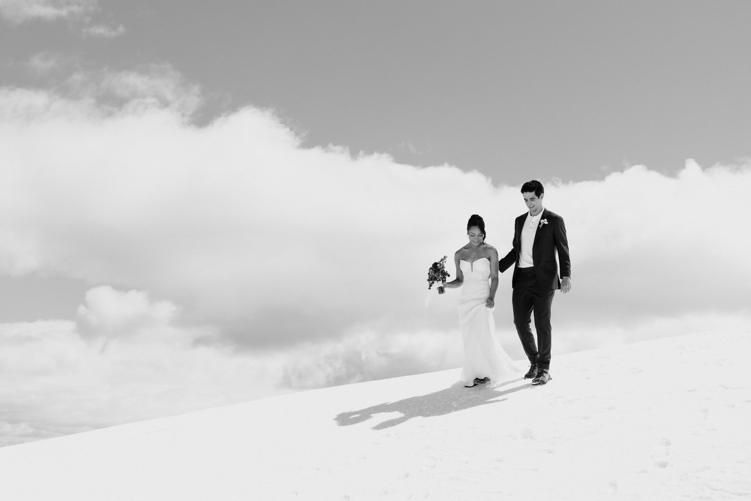 Black and white photograph of a bride and groom walking hand in hand across a snowfield beneath large clouds