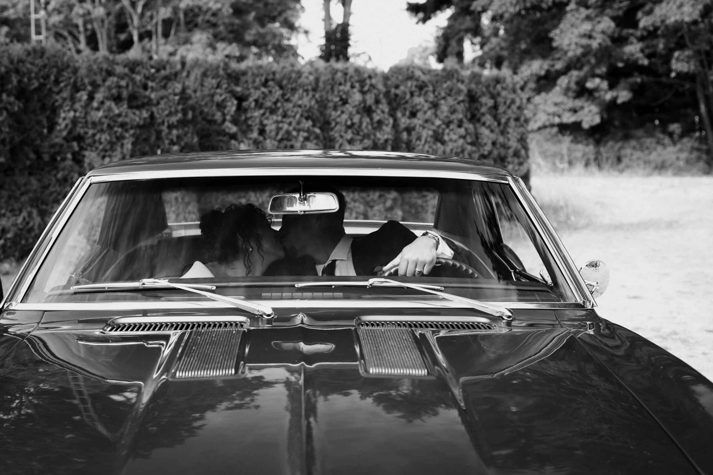 Black and white photograph of a couple kissing, seen through the windscreen of a vintage car