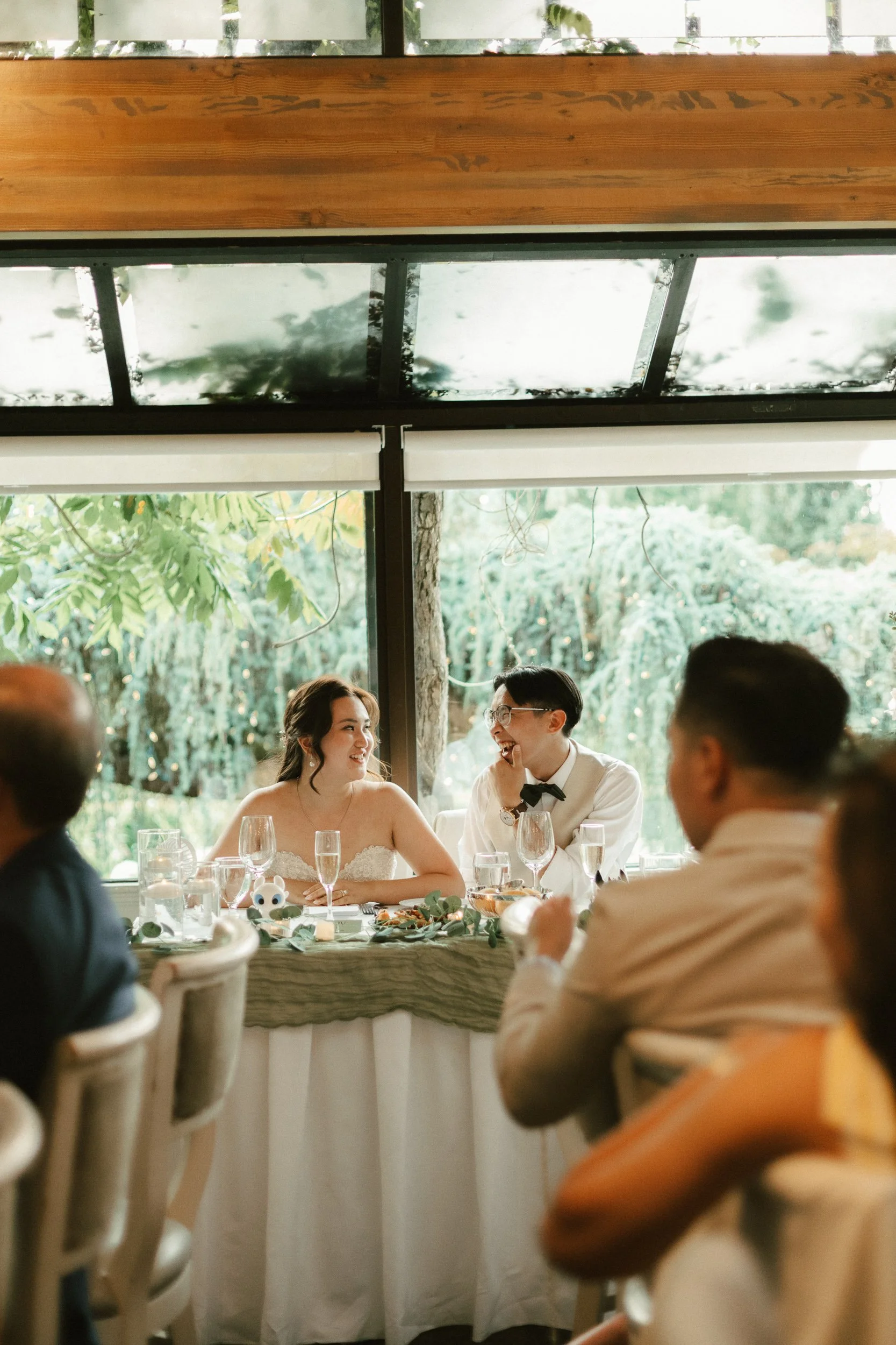 A wedding reception with the bride and groom sitting at a table, smiling and talking, with other guests around them. The scene is seen through a window with greenery outside.