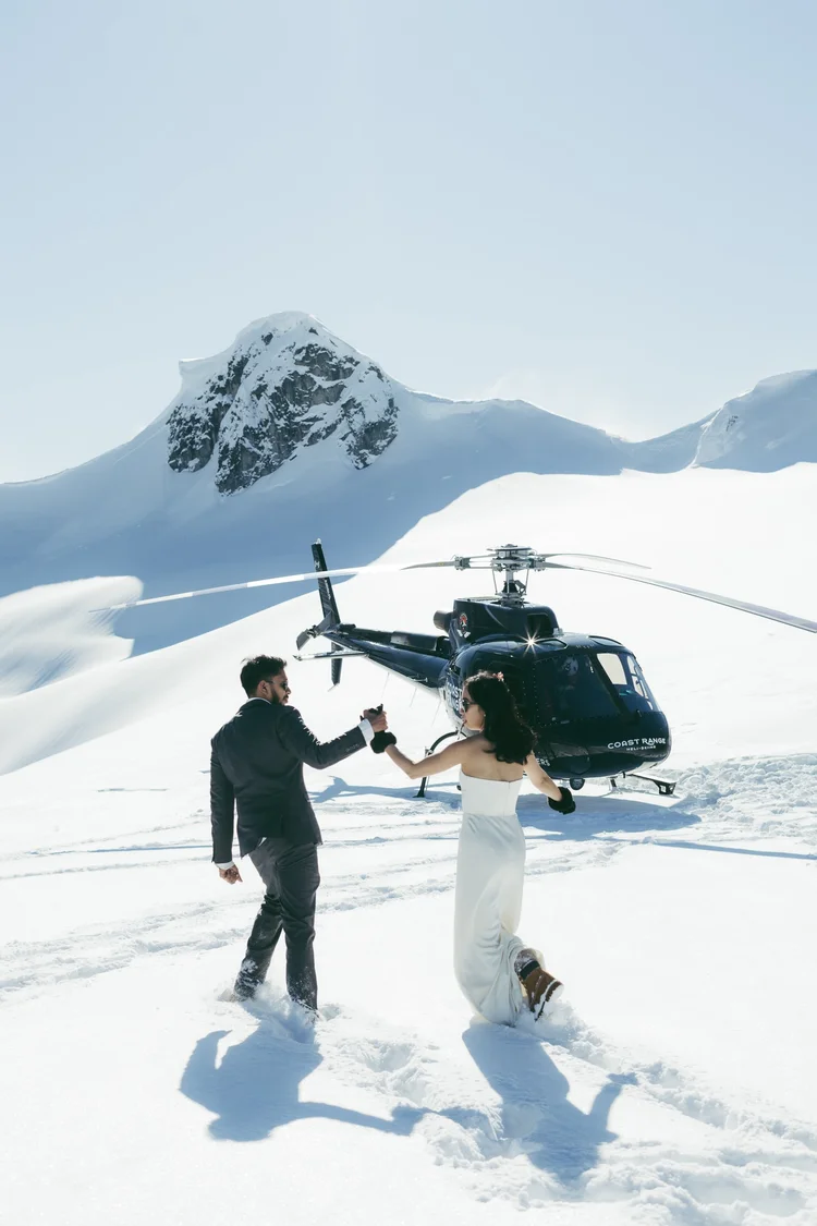 A bride and groom stepping away from a helicopter on a snowy summit beneath a mountain peak