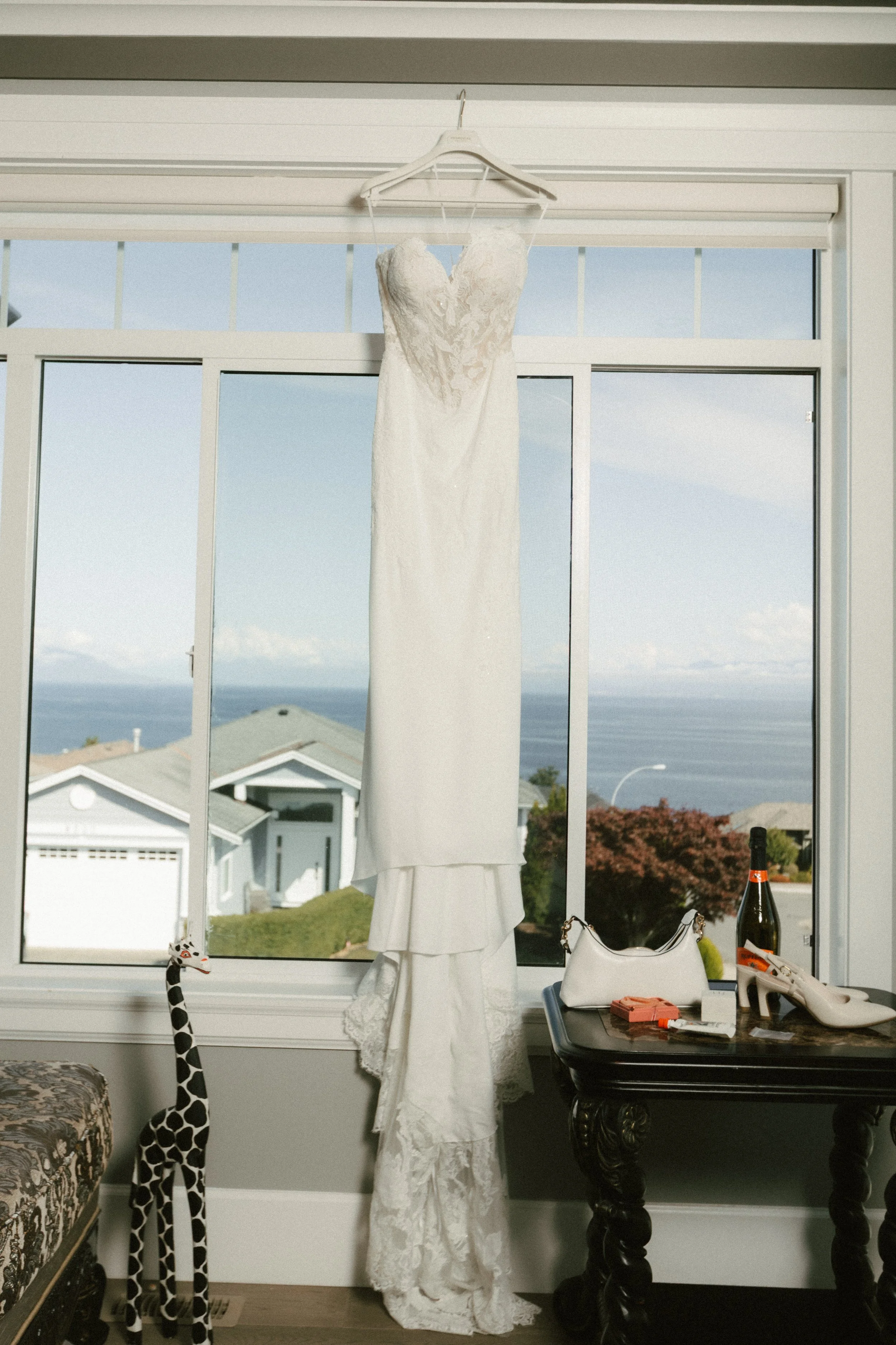 White lace wedding dress hanging in front of a window beside a giraffe sculpture and black console table.