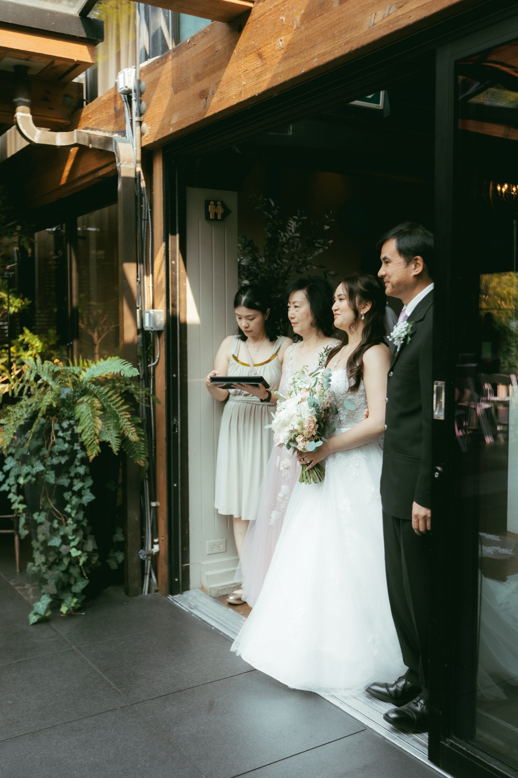 People dressed in formal attire, including a bride in a white wedding gown holding a bouquet, standing with three other women and a man, inside a venue with a wooden ceiling and an open door leading outside. One woman is looking at a tablet.