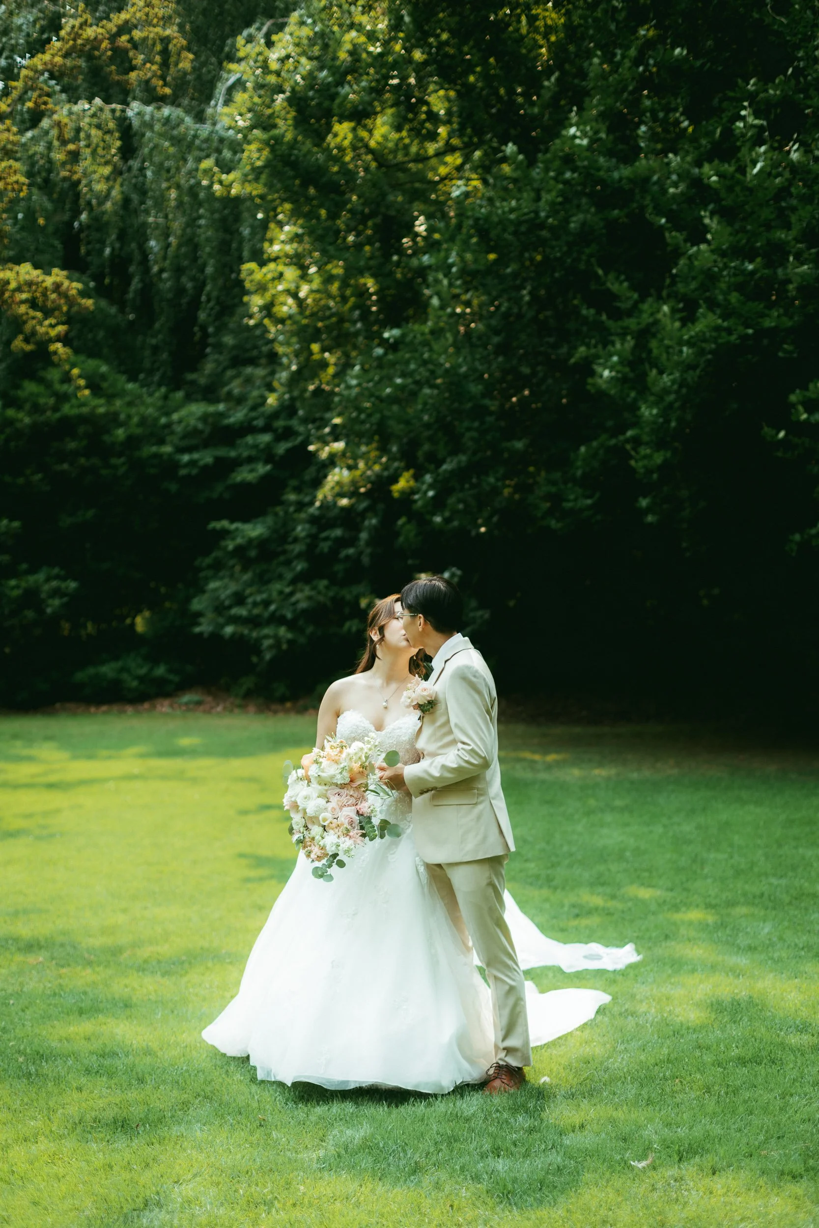 A bride and groom kissing outdoors on a grassy lawn under a background of green trees.