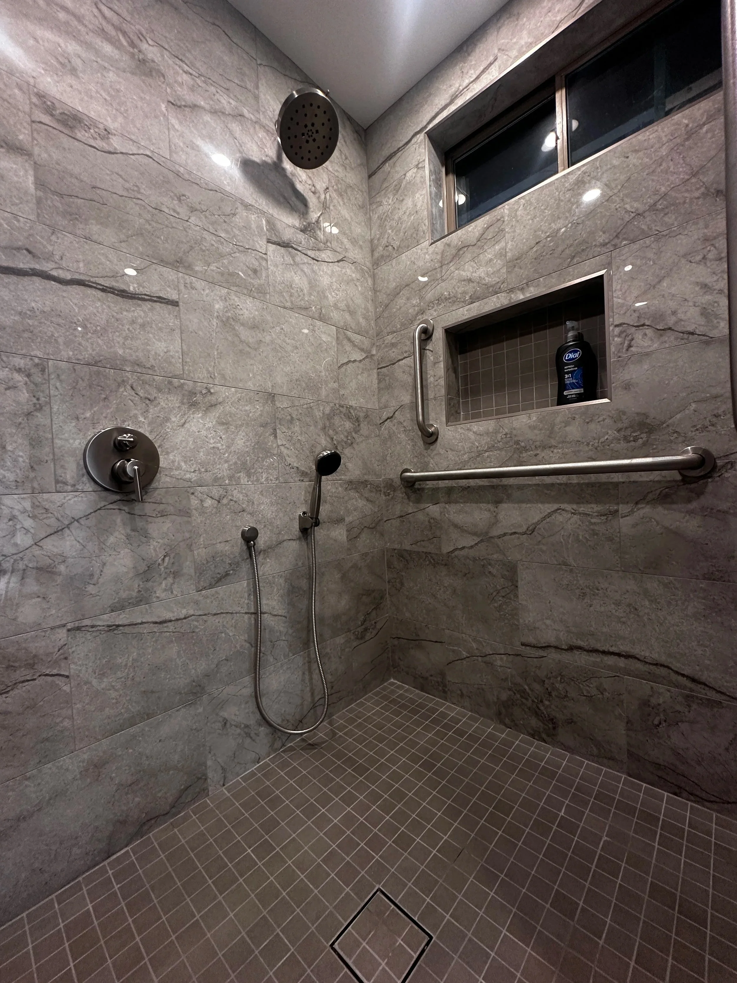 Accessible shower with large gray marbled tiles, a large overhead showerhead, a handheld shower wand, a small window at the top, a niche with shampoo, and grab bars on the wall.