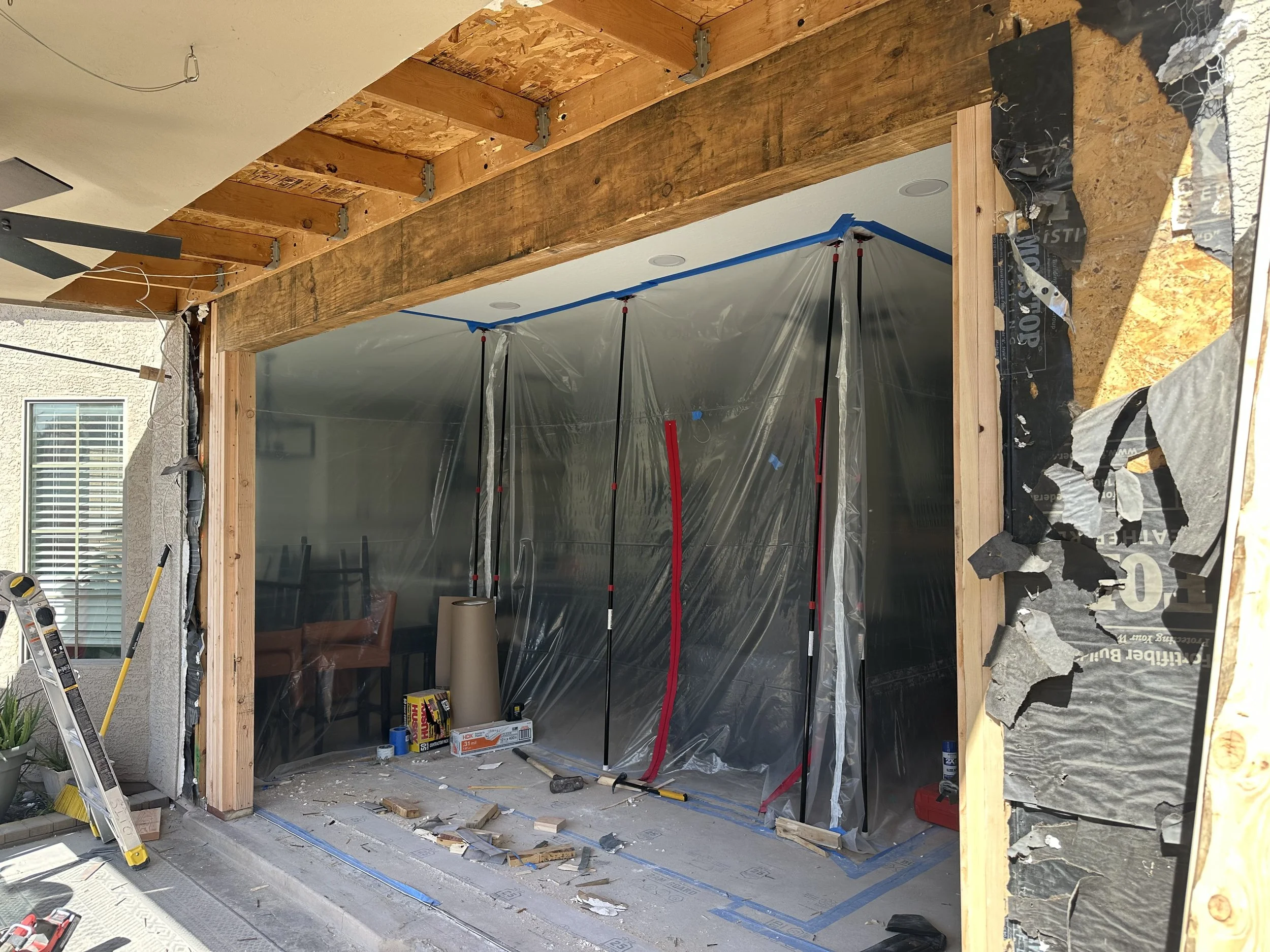Construction site in progress with a partially built wall, plastic sheeting for dust containment, construction tools and materials scattered on the floor, and wooden framing for a new structure.