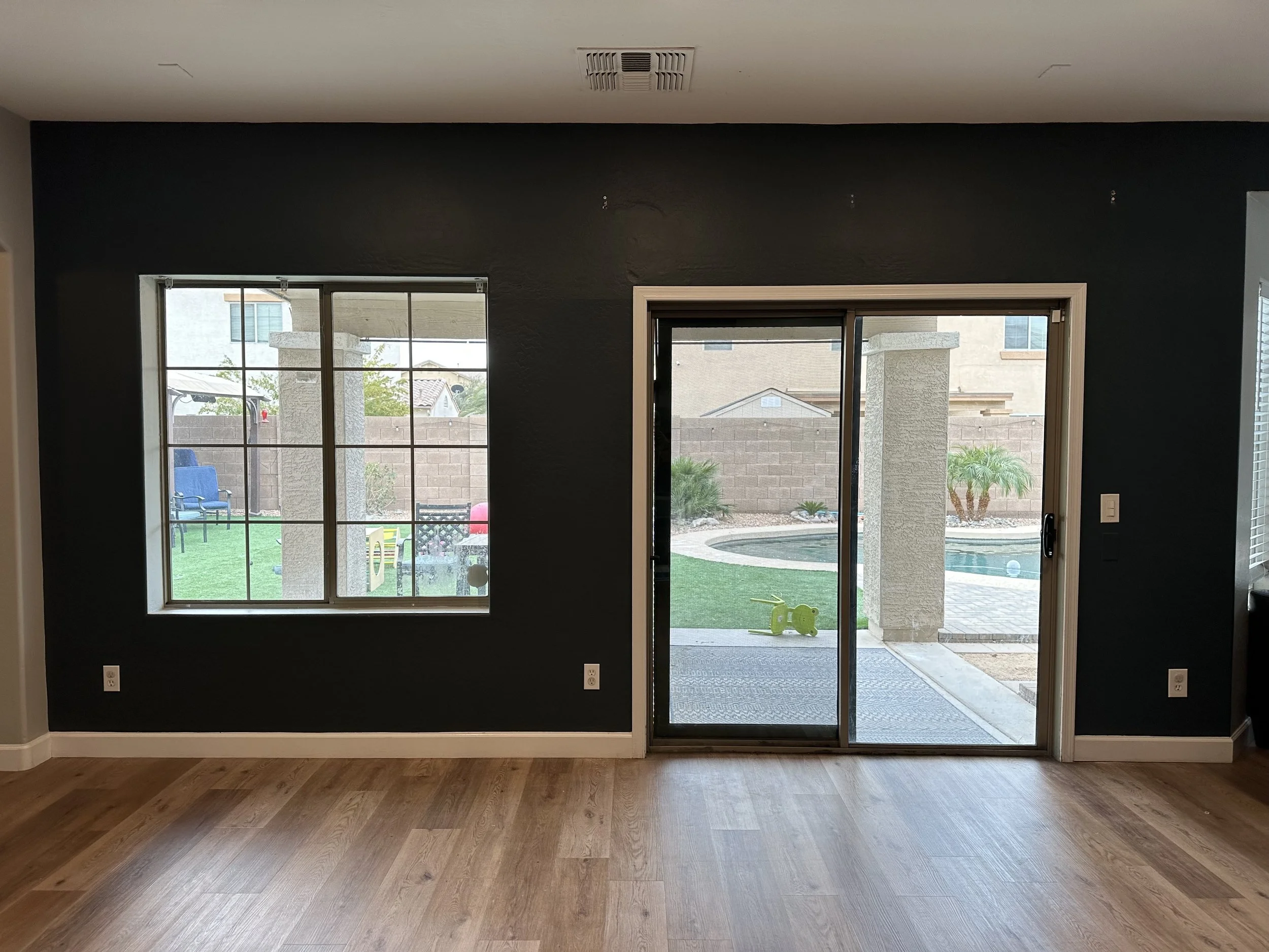 A living room with hardwood flooring, large sliding glass door, and a window, looking out onto a backyard with a pool, patio, and garden.