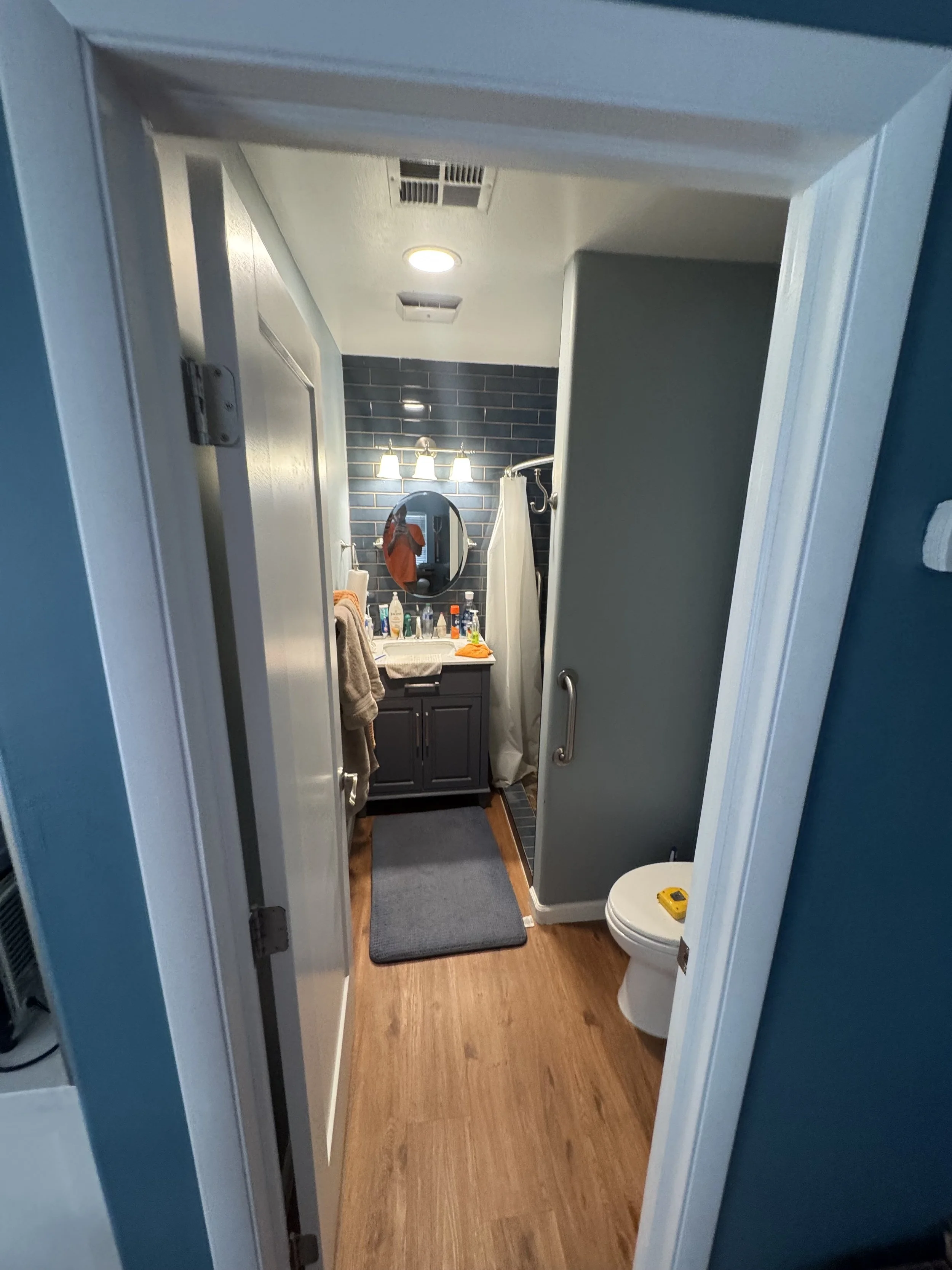 Before picture of a small bathroom with a gray vanity, round mirror, blue tile wall, and shower with curtain before it is remodeled. Bathroom items on the vanity and a gray rug on wooden flooring. Toilet with a tape measure on top.