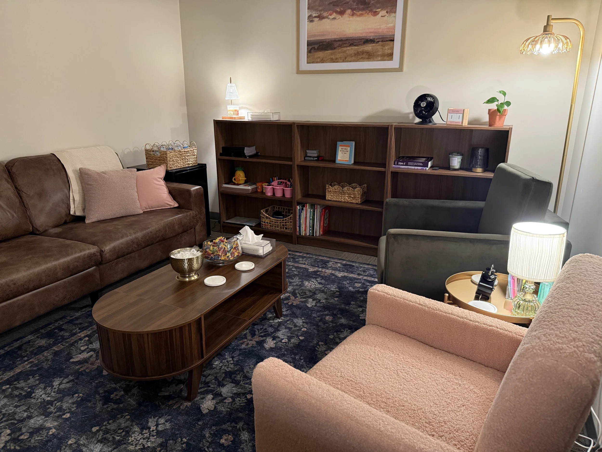 Living room with pink and brown armchairs, a wooden coffee table with candy bowls, a brown sofa with pink and beige pillows, a bookshelf with books and decorations, a lamp, and a framed landscape picture on the wall.