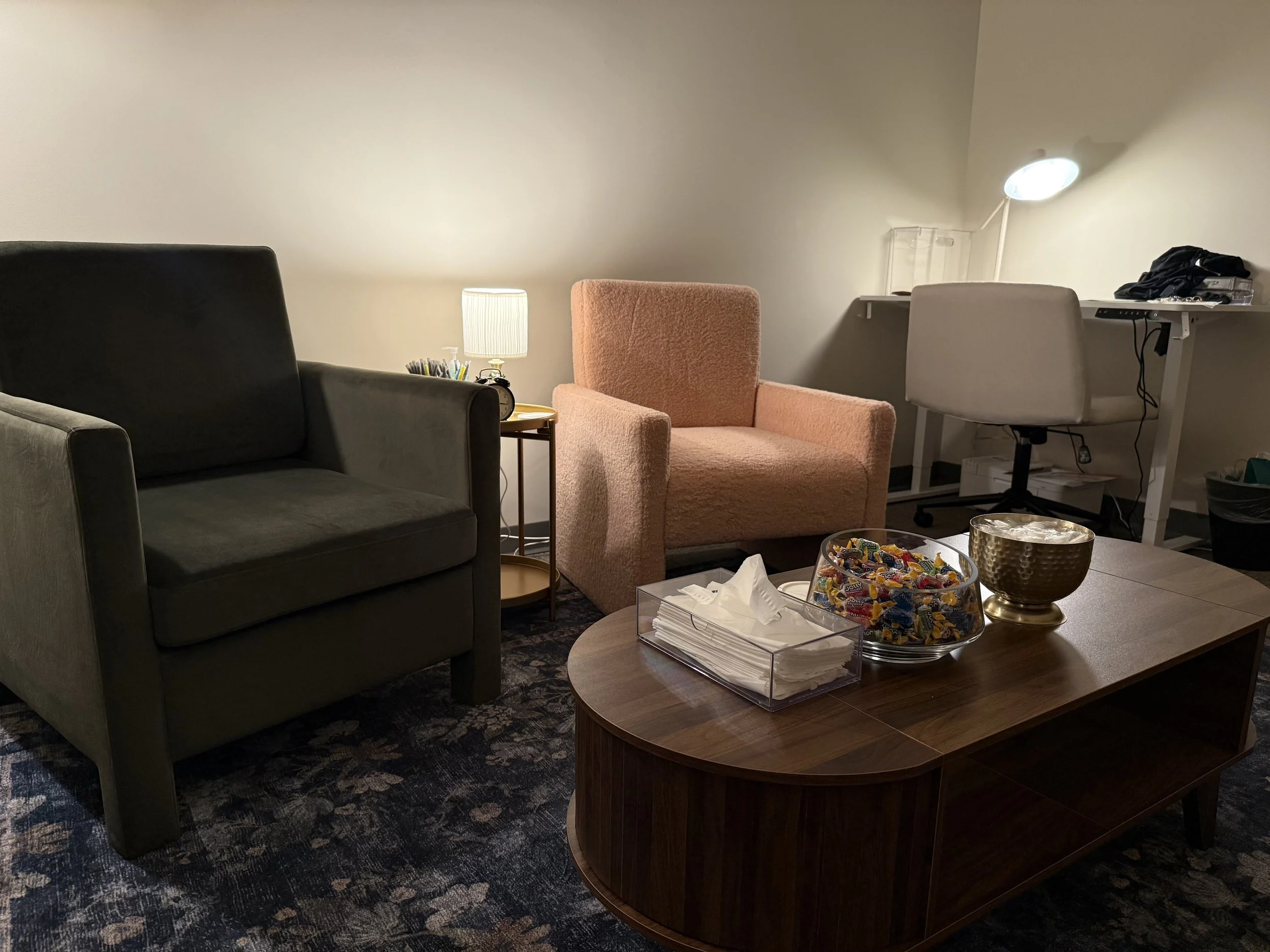 Living room with two armchairs (one black, one pink), a round wooden coffee table with snacks and tissues, a small side table with a lamp, and a white desk with a chair and laptop in the background.