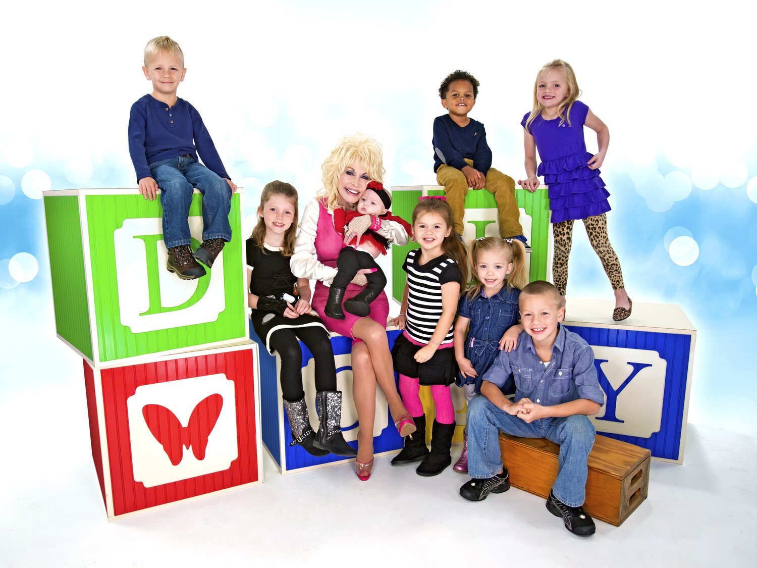 Dolly Parton smiling with a diverse group of children seated around colorful alphabet blocks for her Imagination Library initiative.