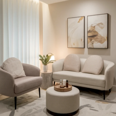A living room with a beige sofa, a matching armchair, a round ottoman with a tray, potted plant, and abstract artwork on the wall.