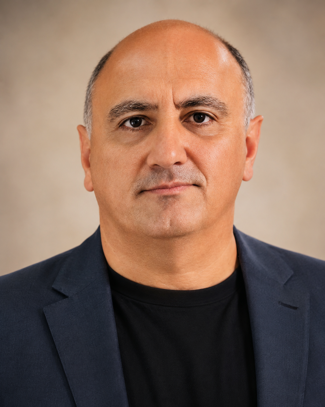 Close-up portrait of a middle-aged man with short gray hair, wearing a dark blazer over a black shirt, looking directly at the camera against a plain background.