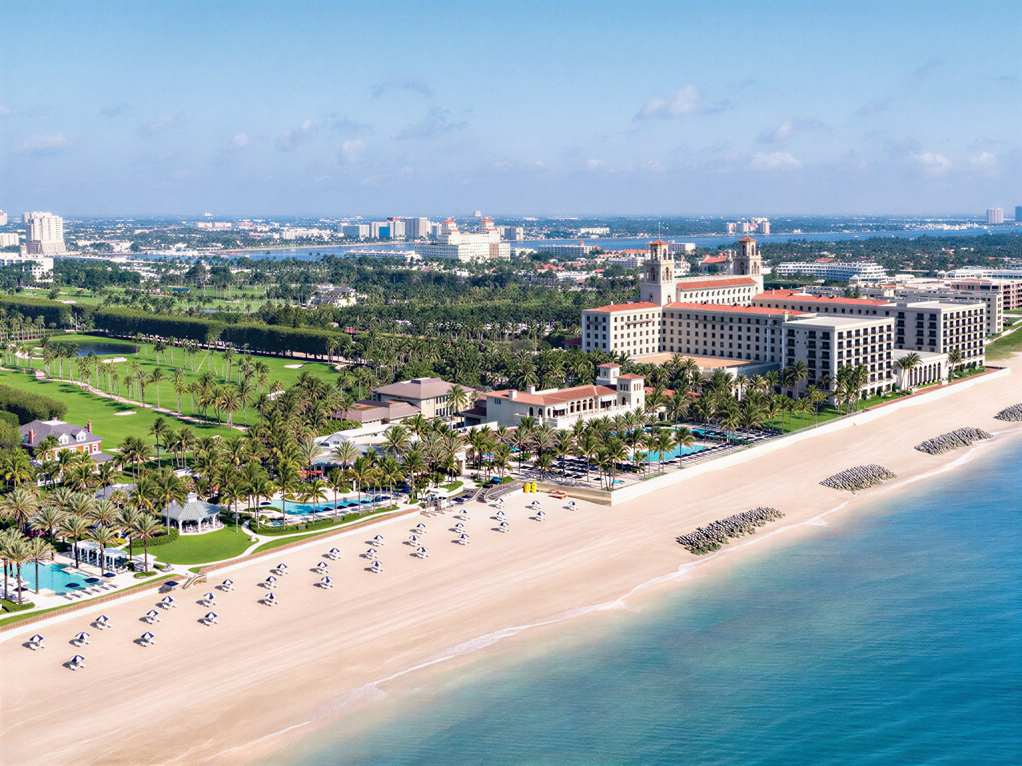 Aerial view of a beach resort with white sand, umbrellas, palm trees, a pool, and a large hotel building near the shoreline. There are city buildings and a river in the background under a partly cloudy sky.
