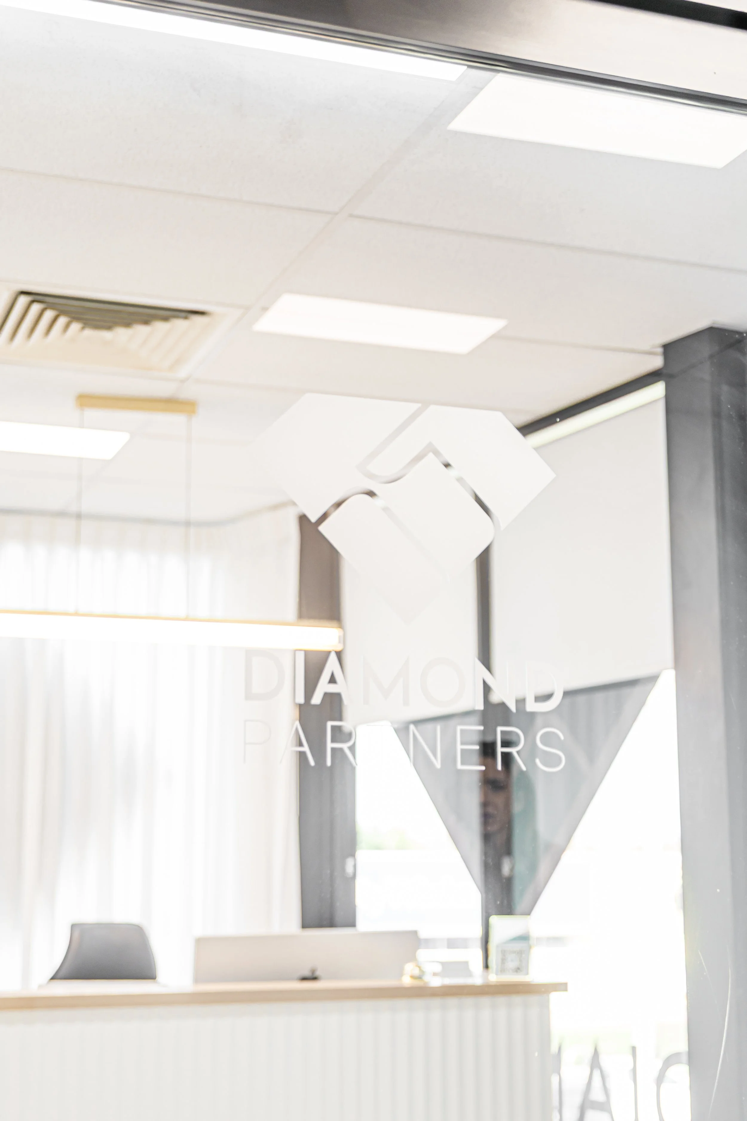 Interior office reception desk with the name 'Diamond Partners' on a glass panel, with a white ceiling, fluorescent lights, and a person partially visible behind the glass.