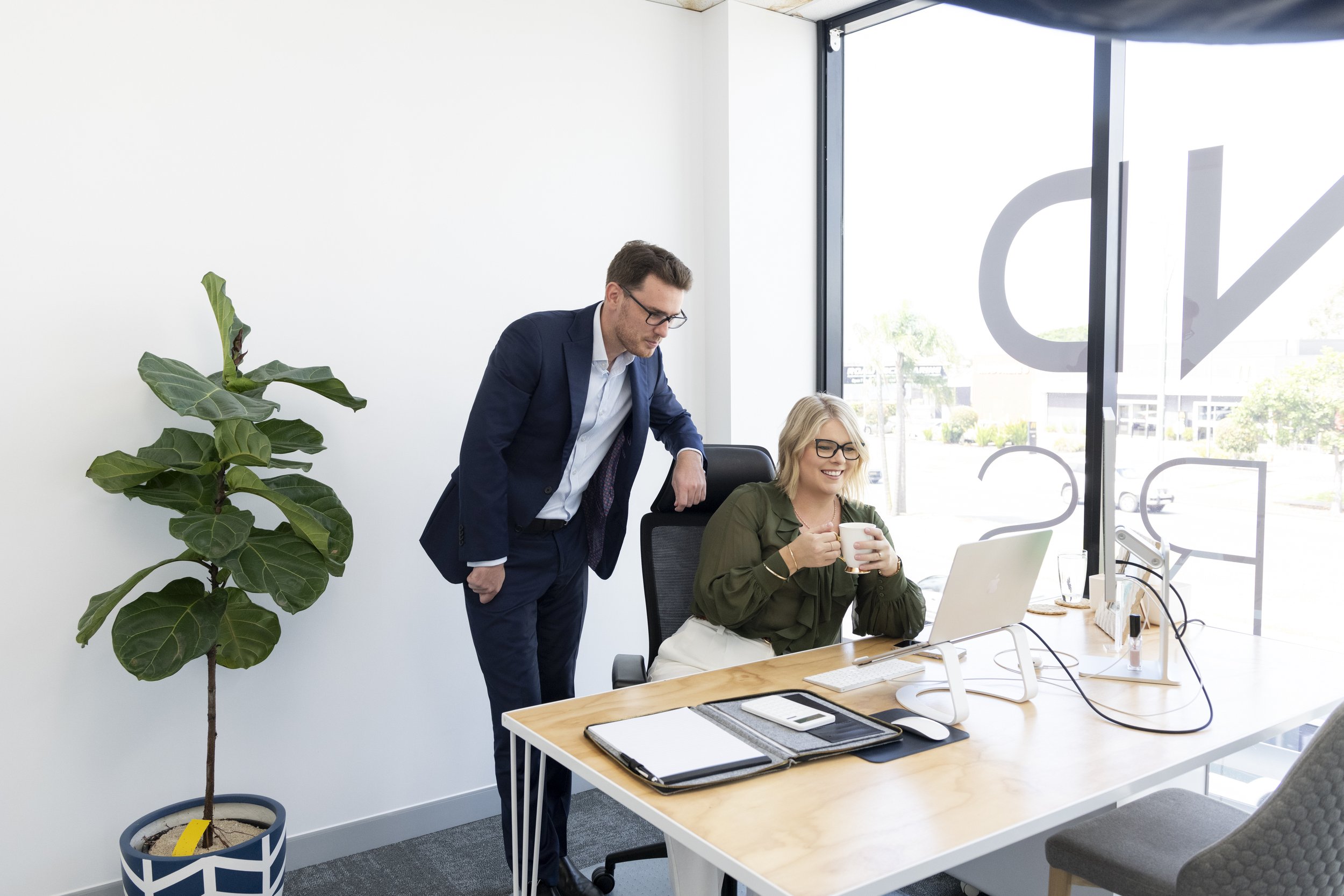 A man and woman in an office, with the man leaning over the woman who is sitting at a desk holding a mug, laptop, and office supplies, near a large window and a potted plant.