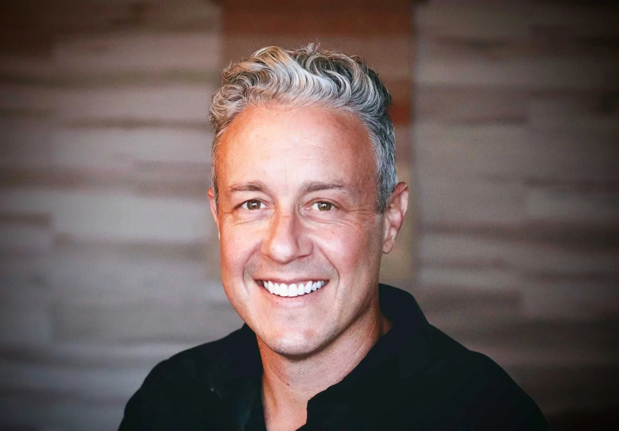 A smiling middle-aged man with gray and white hair, wearing a black shirt, against a blurred wooden background.