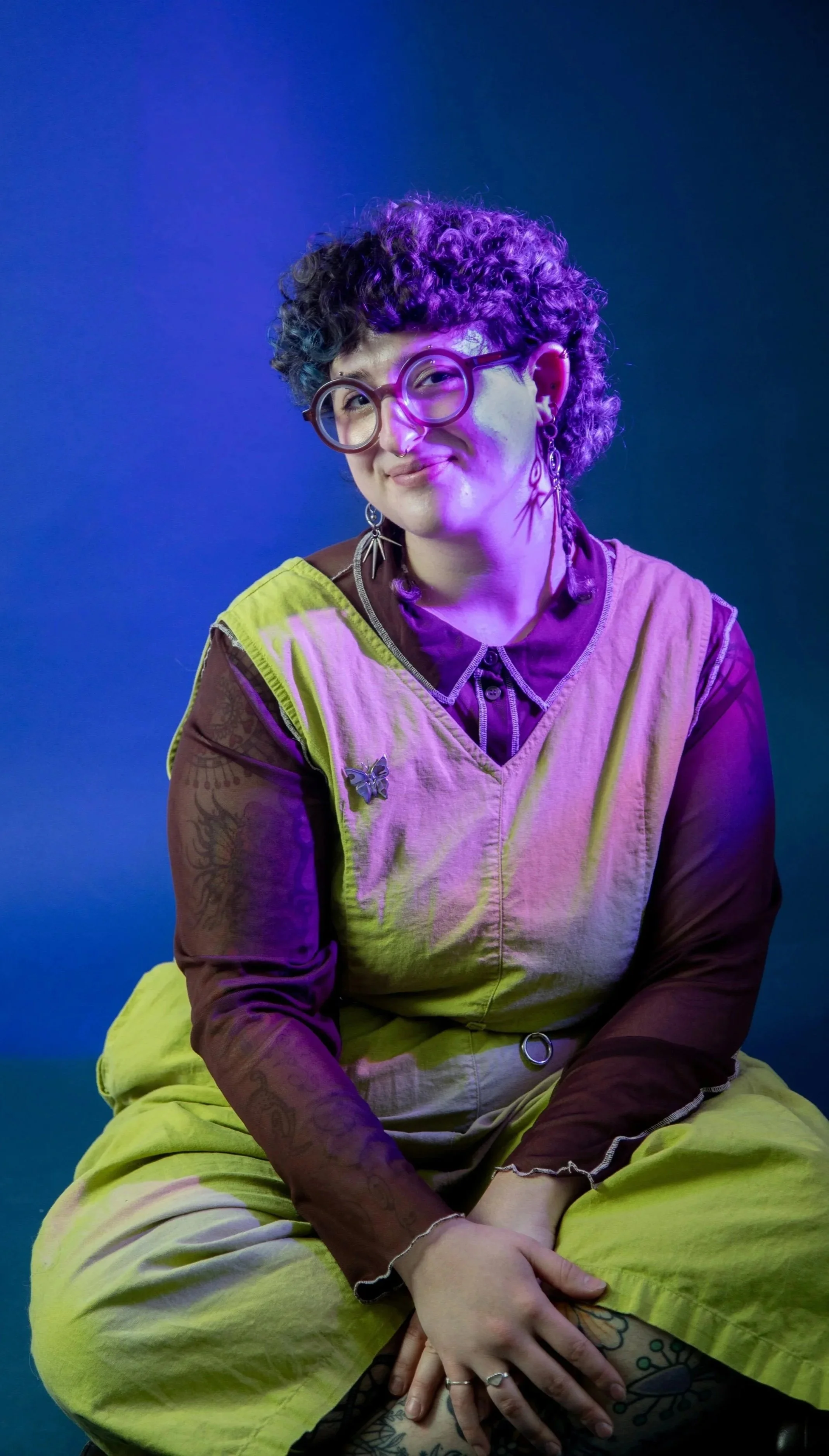 A smiling person, seated on the ground, wearing a green jumpsuit and brown button-up shirt.