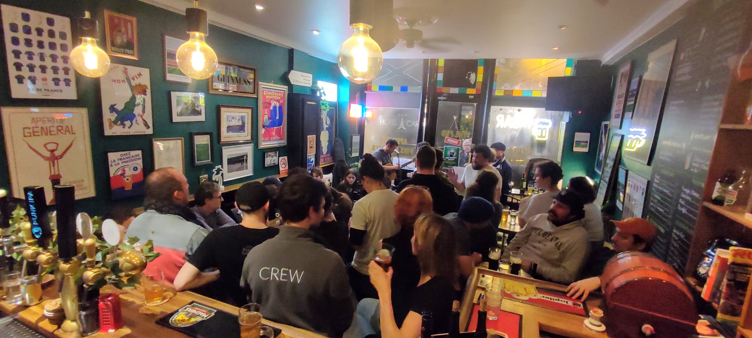 A crowded bar with people socializing, drinking, and enjoying live music in a cozy, decorated space with framed pictures and neon signs.