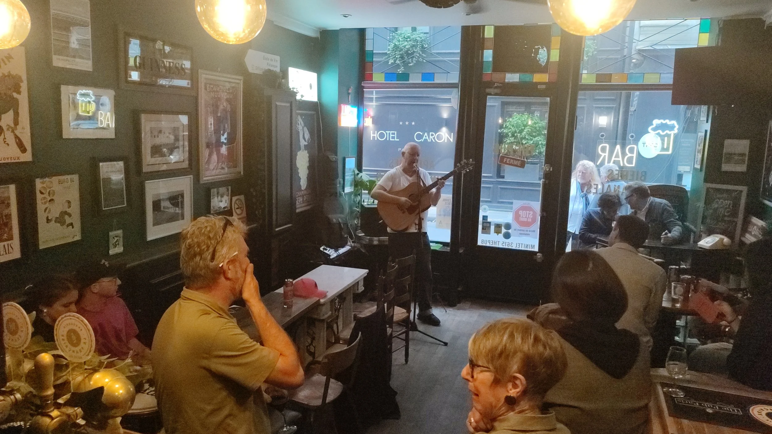 Man playing guitar and singing in a pub with an audience seated around tables, some looking toward the performer. Window with neon signs and outdoor seating visible.