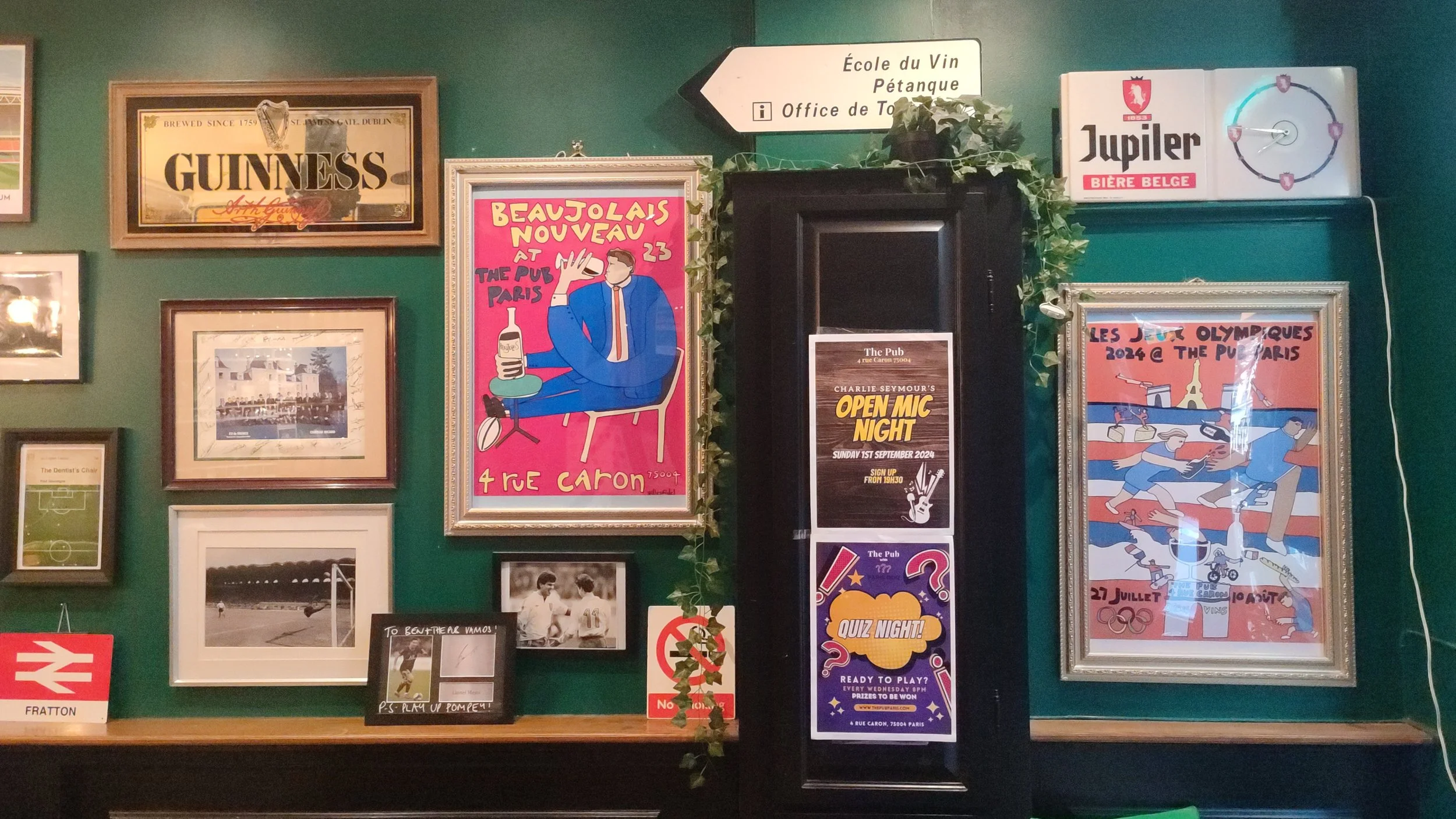 Decorative wall with framed posters, photographs, and signs at a pub. Posters include a colorful illustrated promotional for Beaujolais Nouveau, a black and white photo of a soccer goal, and a poster for Olympic diving.