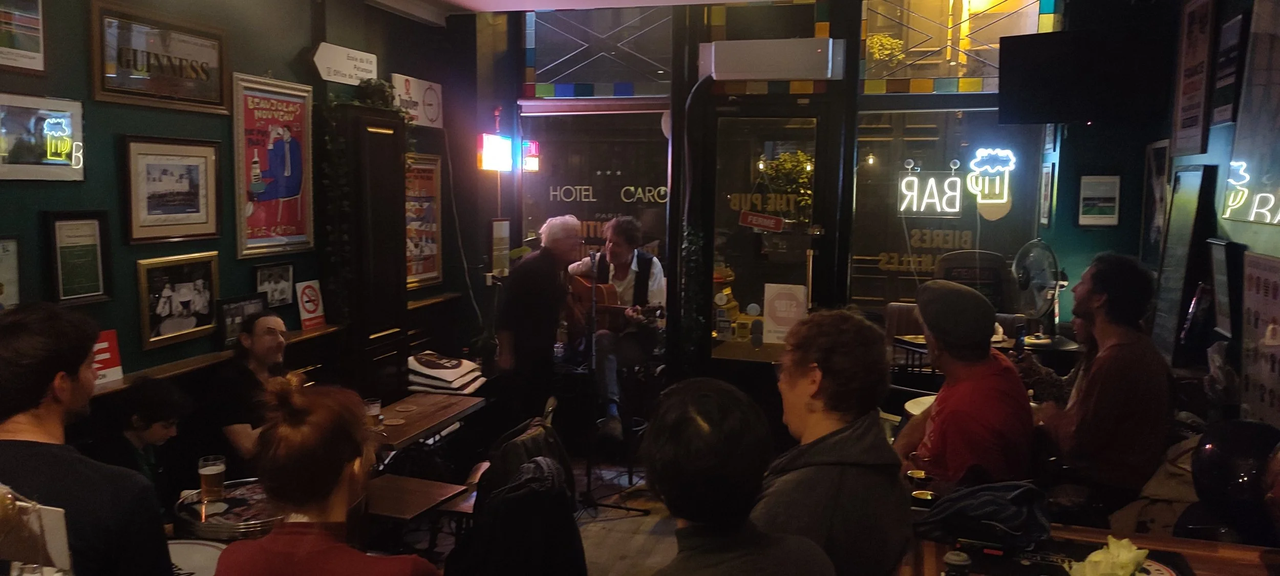 People gathered in a cozy pub, enjoying live acoustic music performance. The scene features framed pictures on the green walls, neon signs, and a large window revealing the street outside.