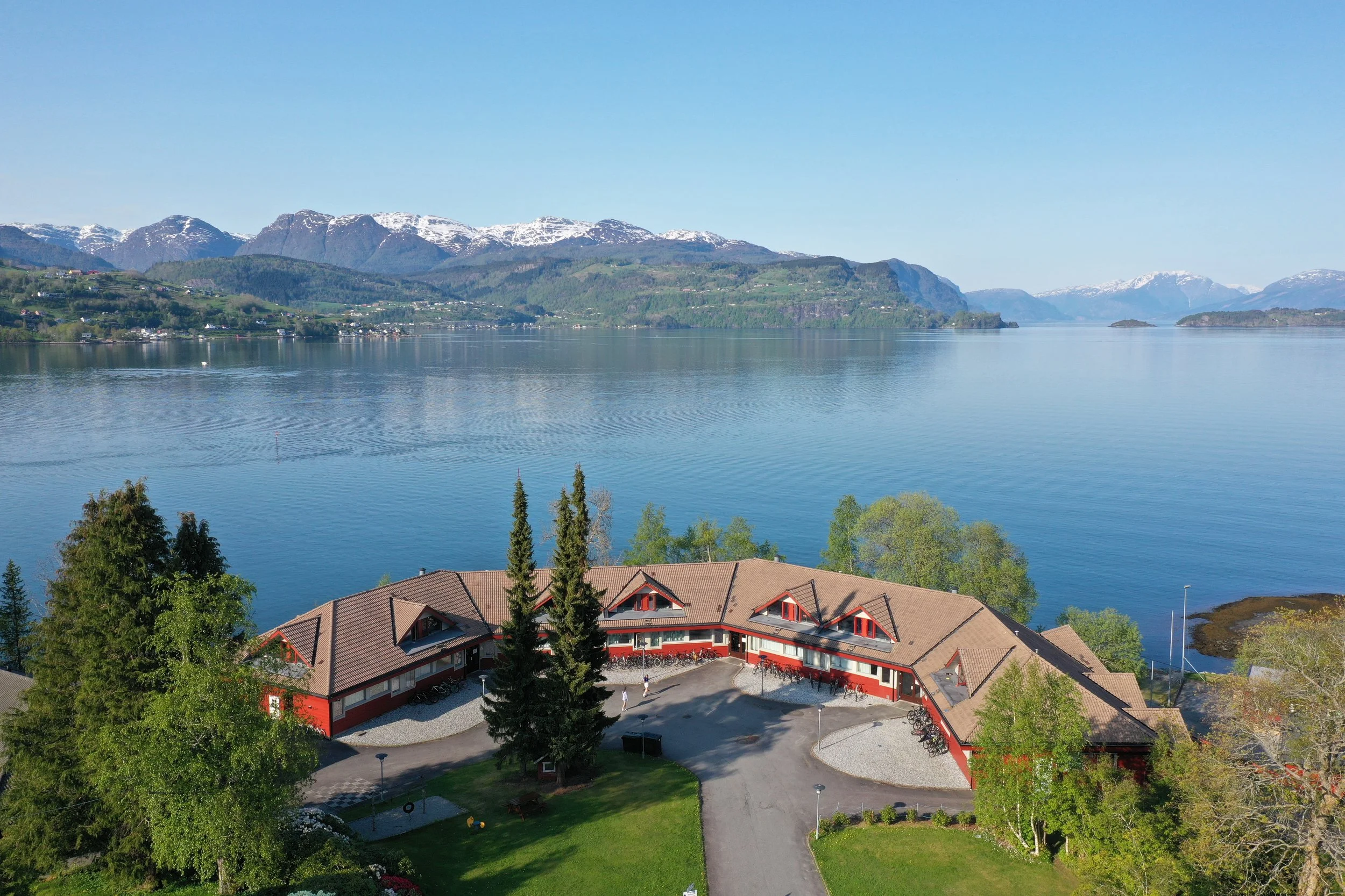 Raudt internatbygg ved Hardangerfjorden, med snødekte fjell på andre sida av fjorden