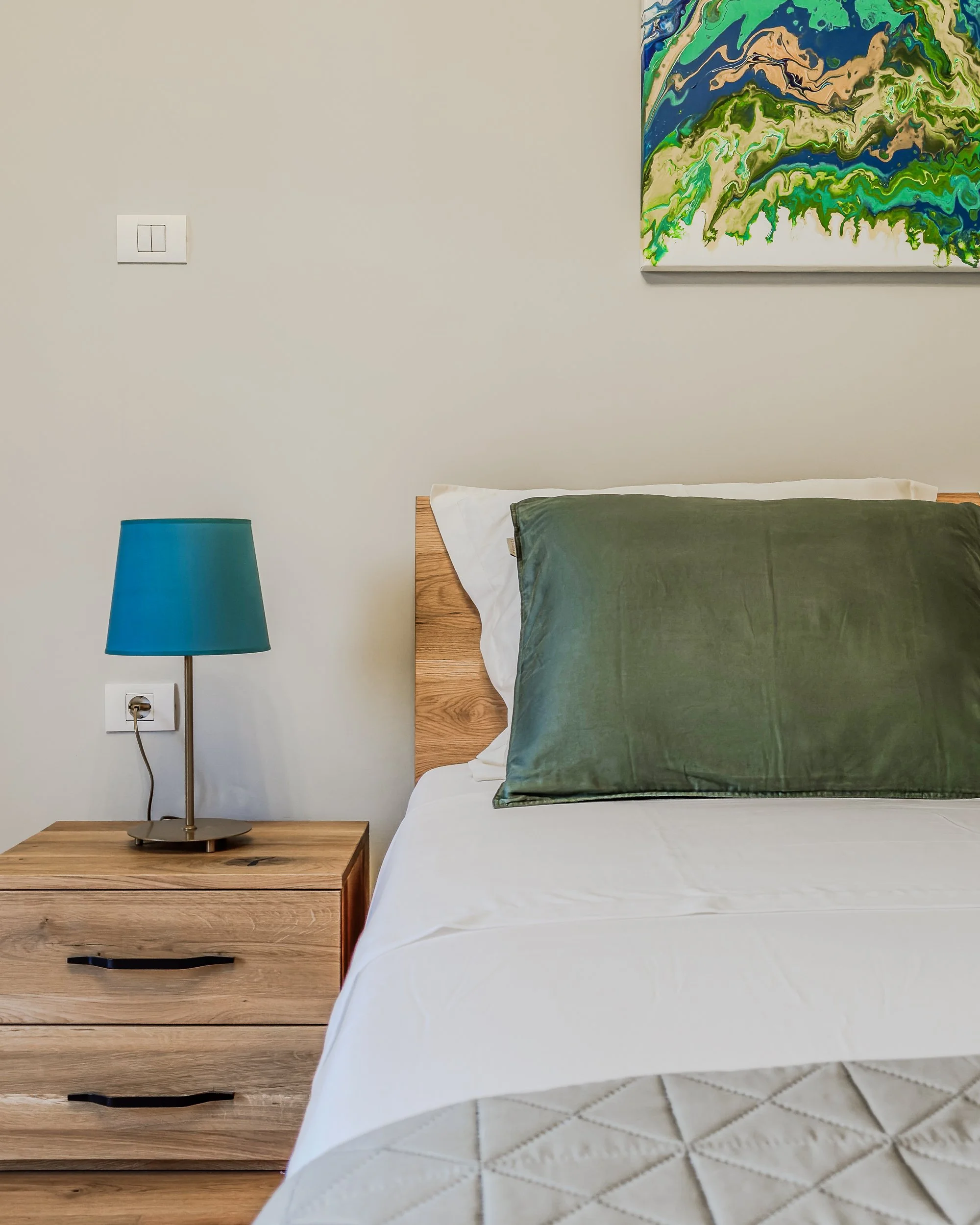 A bedroom with a wooden nightstand, a teal bedside lamp, and a bed with a green pillow, white sheets, and a white quilt. There is an abstract painting with blue, green, beige, and black colors on the wall above the bed.