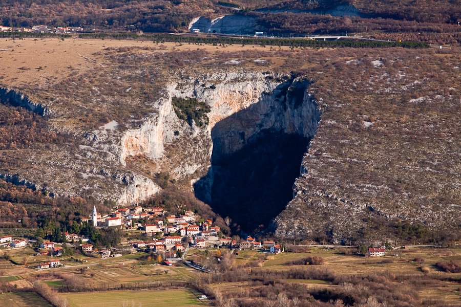 A small village at the base of a large, rocky cliff with a deep, dark cave opening in the middle.