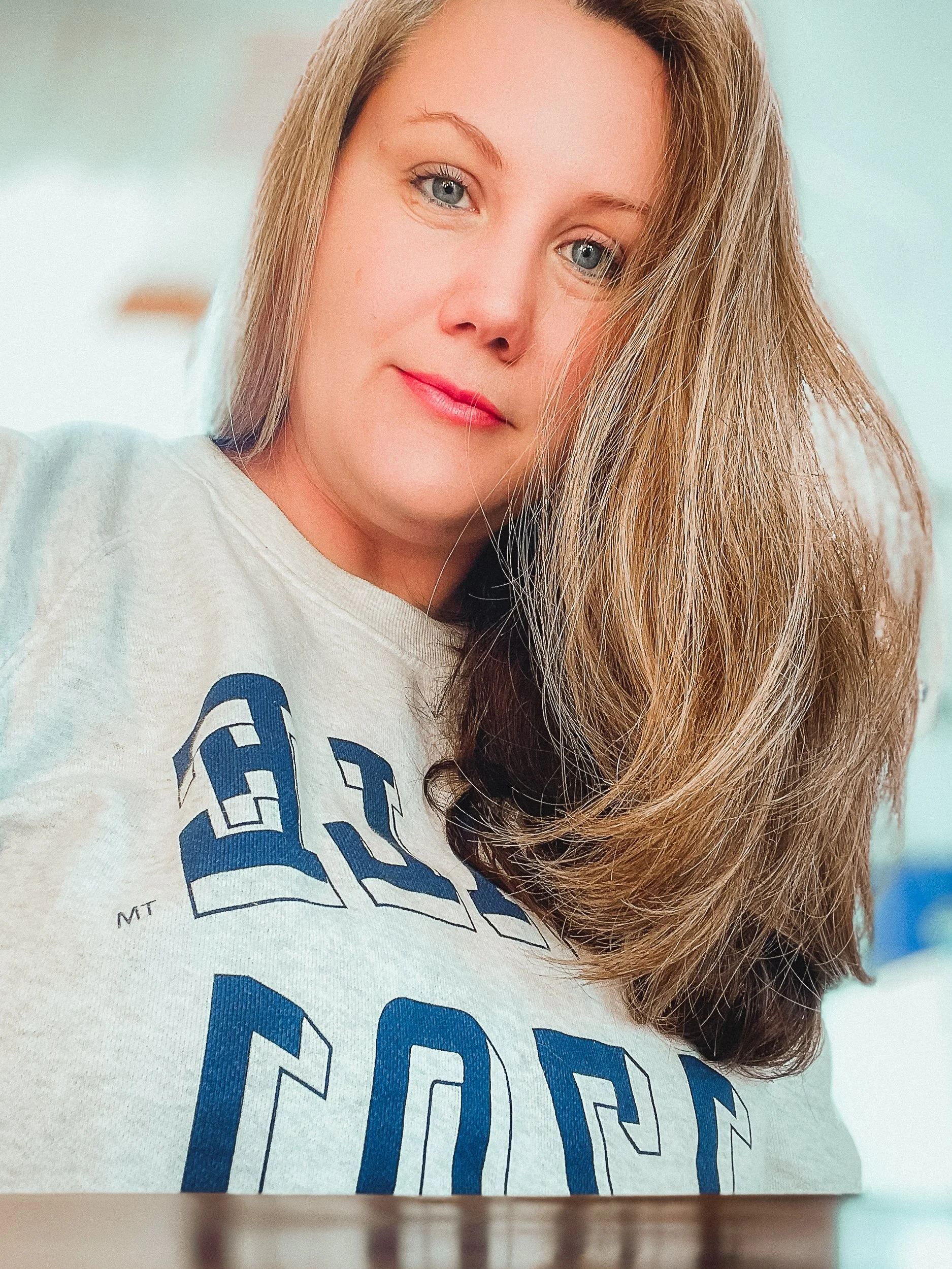 A woman with long, wavy light brown hair and blue eyes looking at the camera, wearing a gray sweatshirt with blue text.