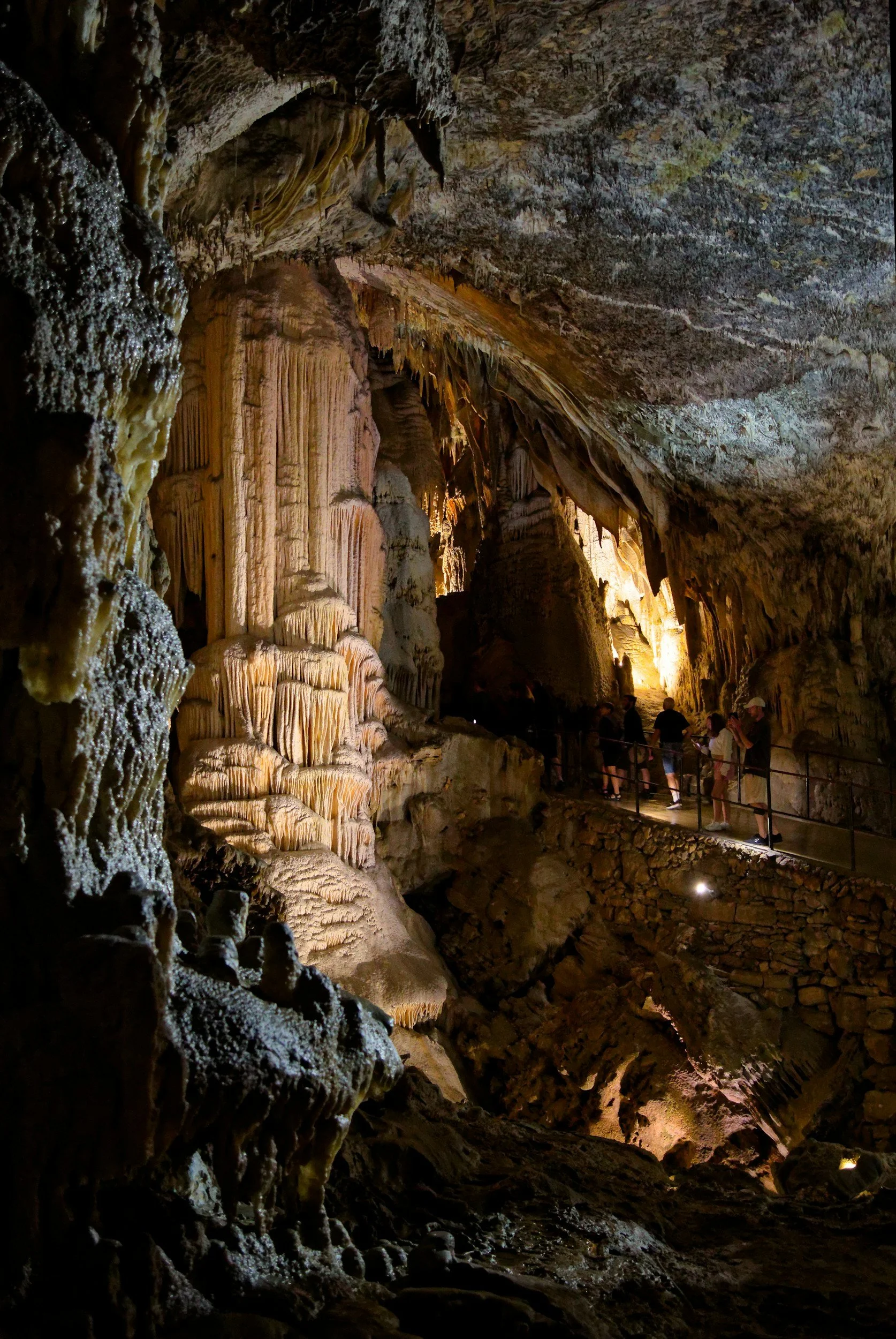 Inside a large limestone cave with stalactites and stalagmites, with a group of people walking along a narrow pathway.