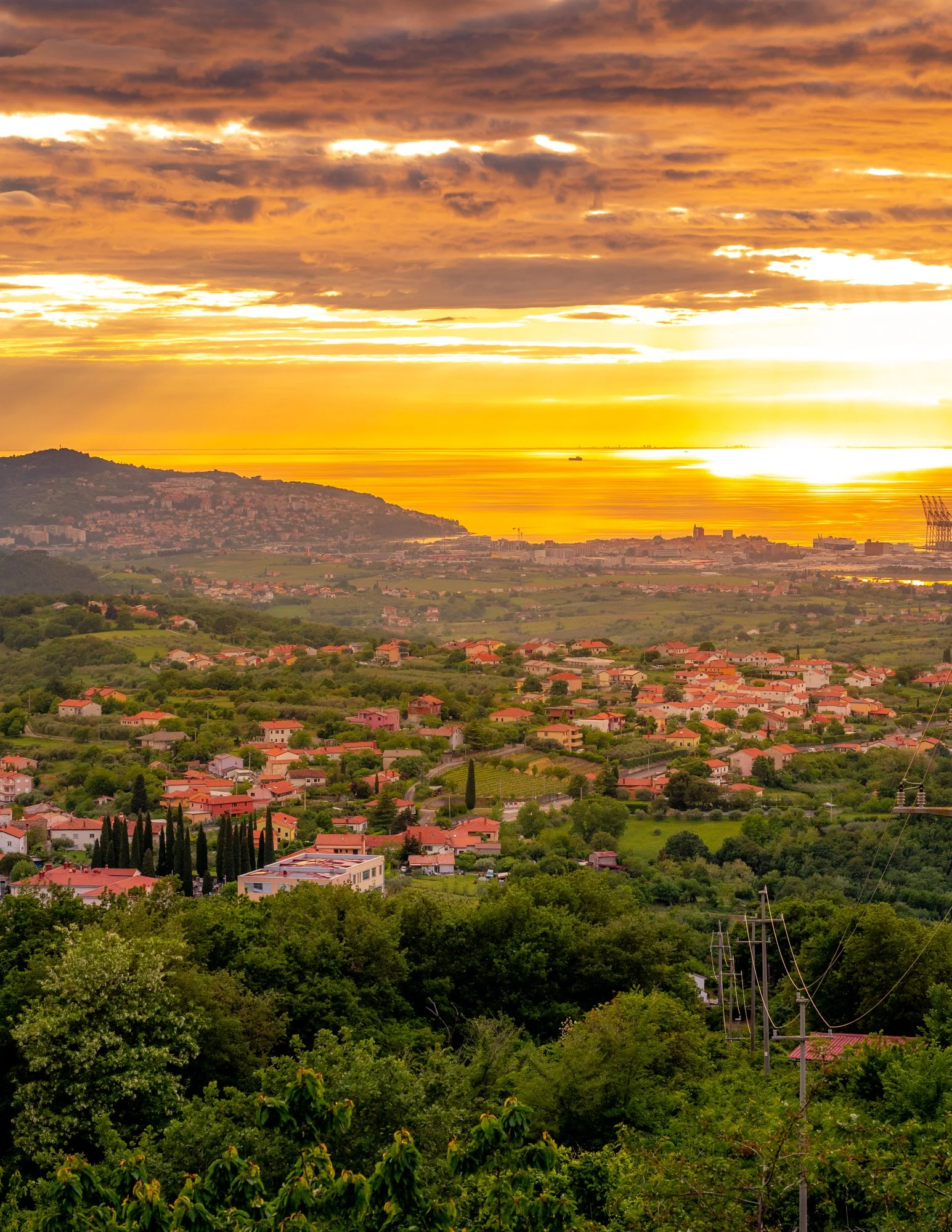 A scenic view of a suburban landscape at sunset, with green trees, houses, and hills in the background, and a bright sky with orange, yellow, and dark clouds.