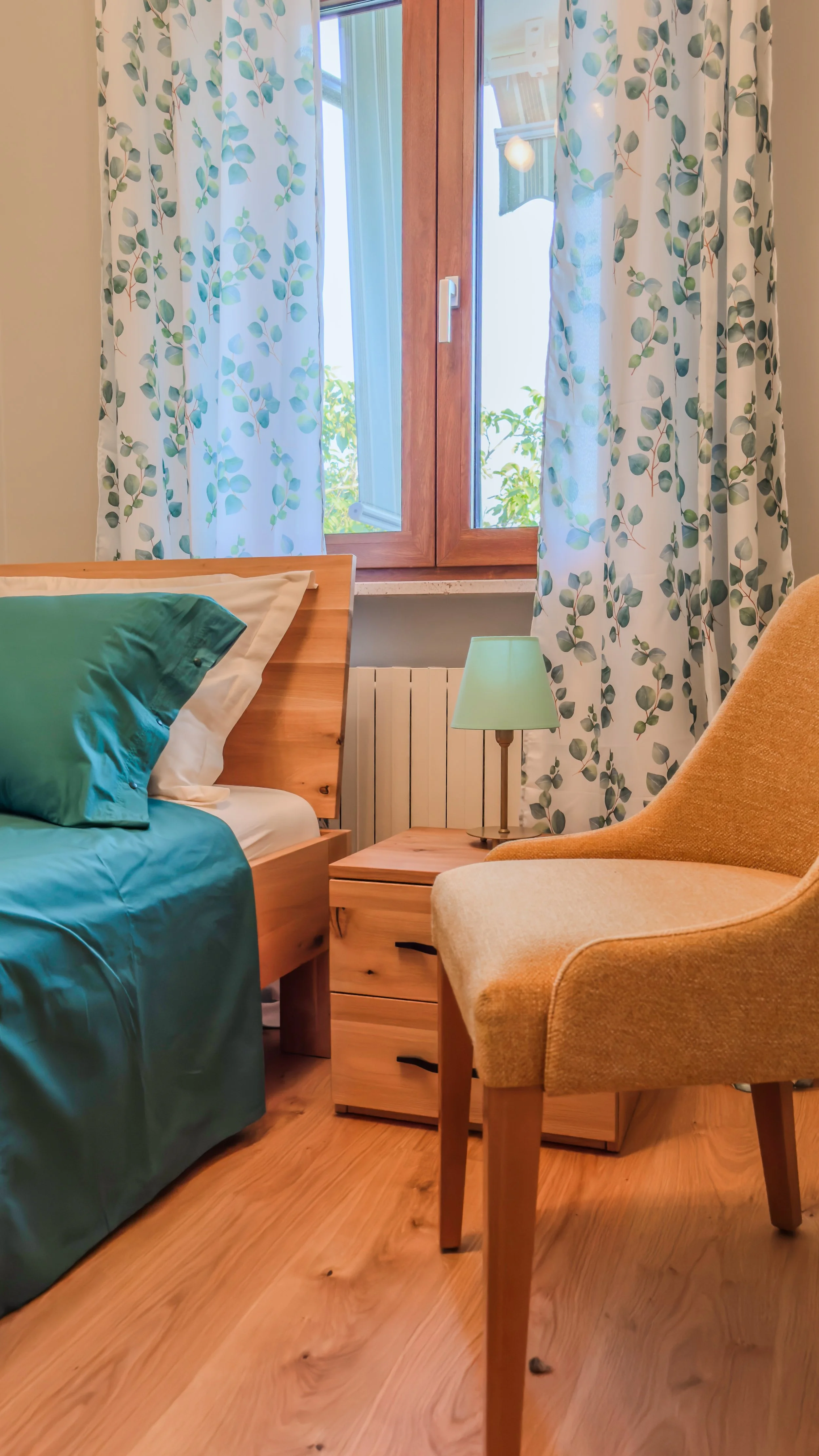 A bedroom corner with a wooden bed, green bedding, a wooden nightstand with a green lamp, an upholstered beige chair, and a window with floral curtains, sunlight outside. Located at Villa Domenico.