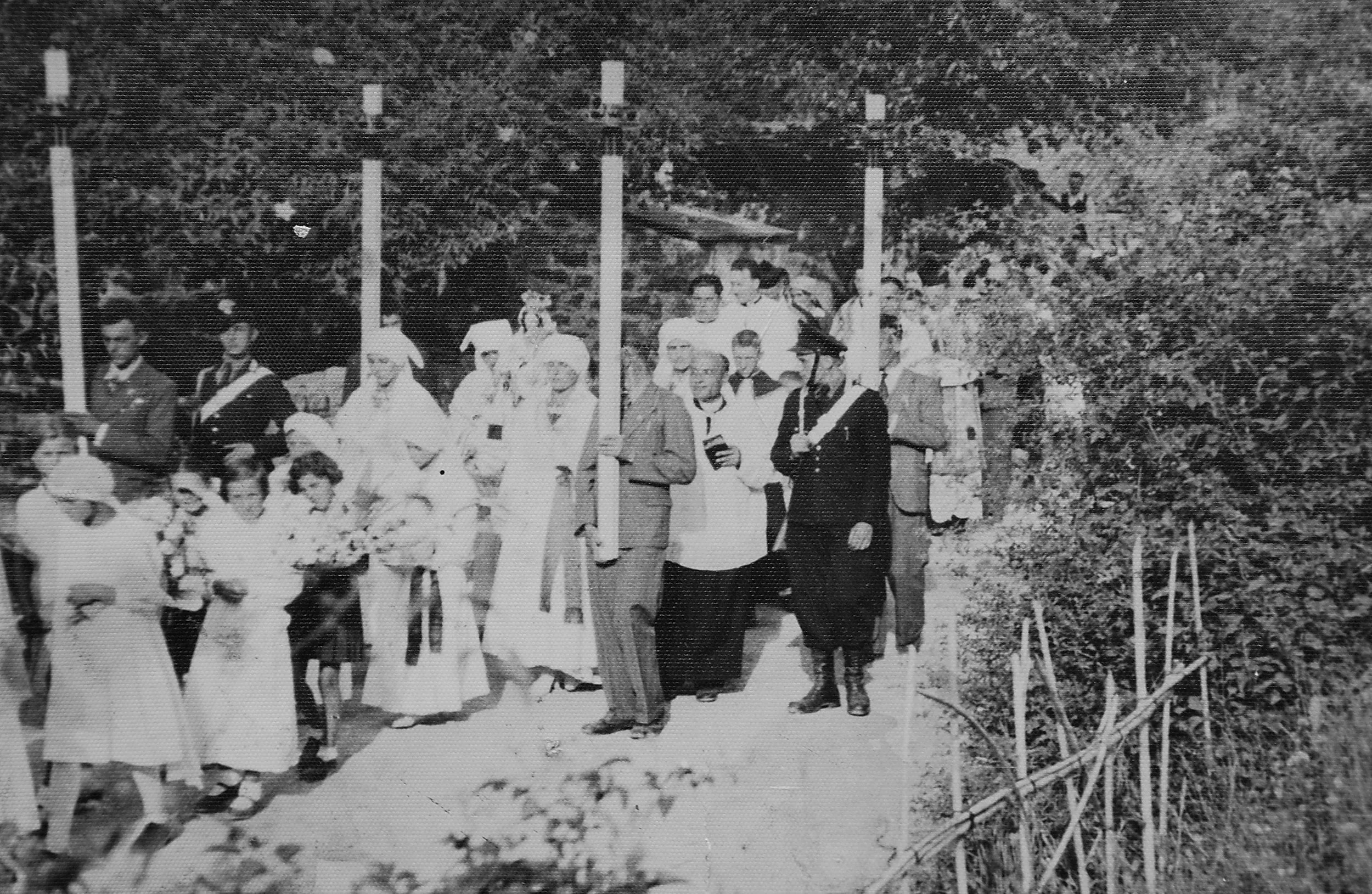 Black and white photograph of a group of people, including women, men, and children, standing outdoors among trees. Some individuals are dressed in formal or traditional clothing, and a few are holding objects. The setting appears to be a garden or park with foliage and a pathway.