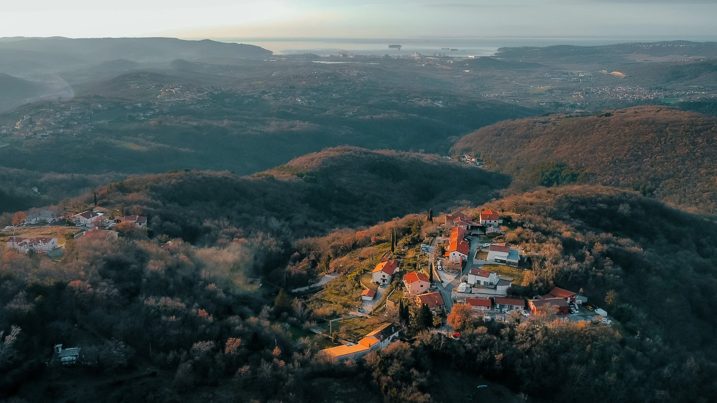 Aerial view of a small hillside neighborhood with houses and trees, surrounded by rolling green hills and a distant view of the water and ships.