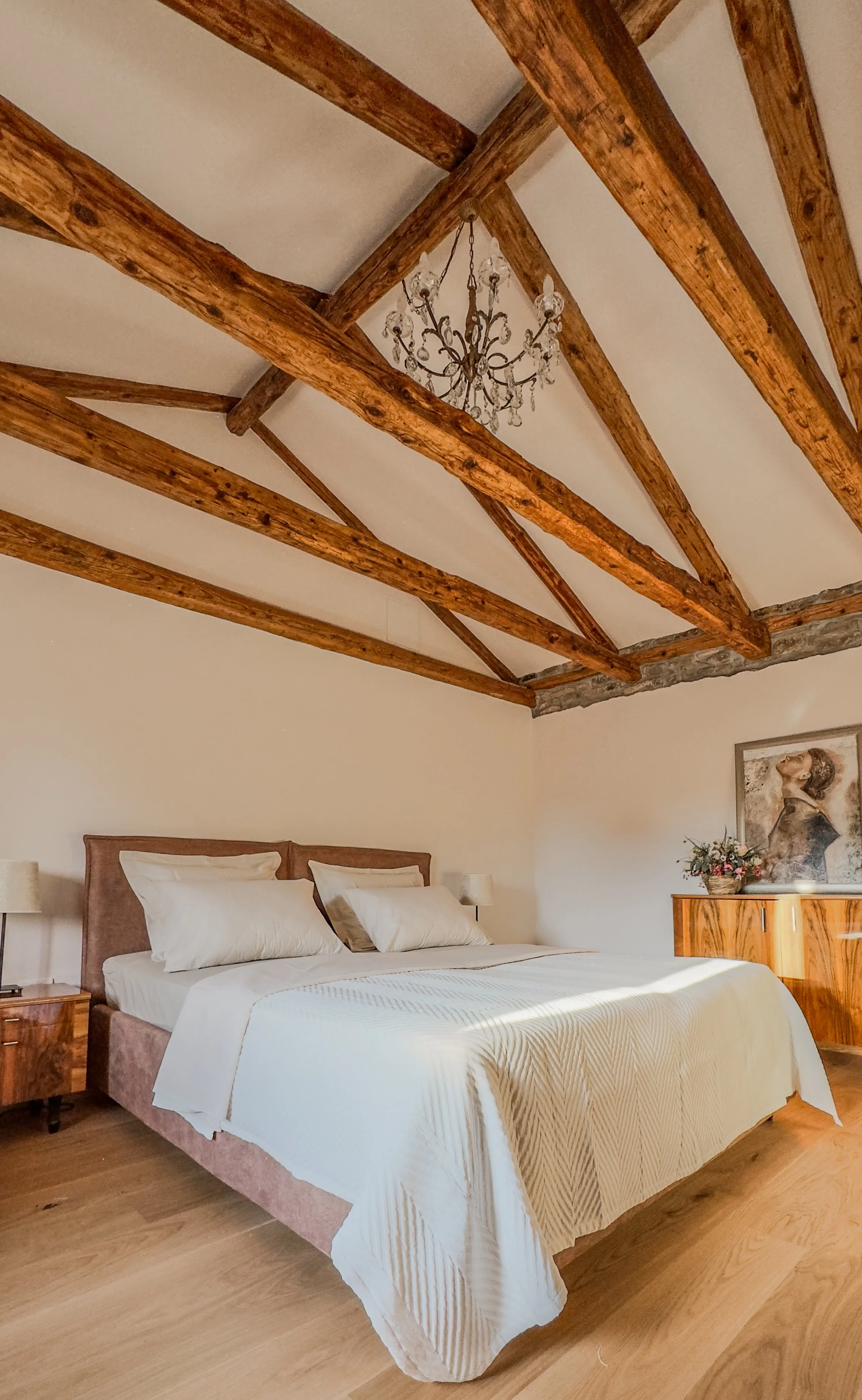 A bedroom with a king-sized bed, wooden nightstand with a lamp, wooden dresser with a flowerpot and a framed portrait, vaulted ceiling with exposed wooden beams, and a chandelier hanging from the ceiling.