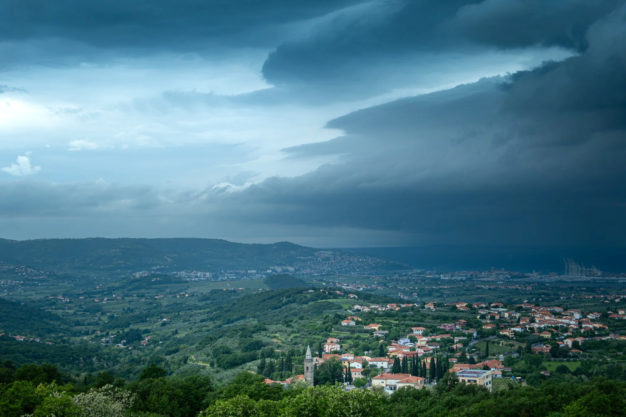 Scenic view of a lush green landscape with a small town, hills, and a coastline under a dark, cloudy sky. Photo: Natali Gak.