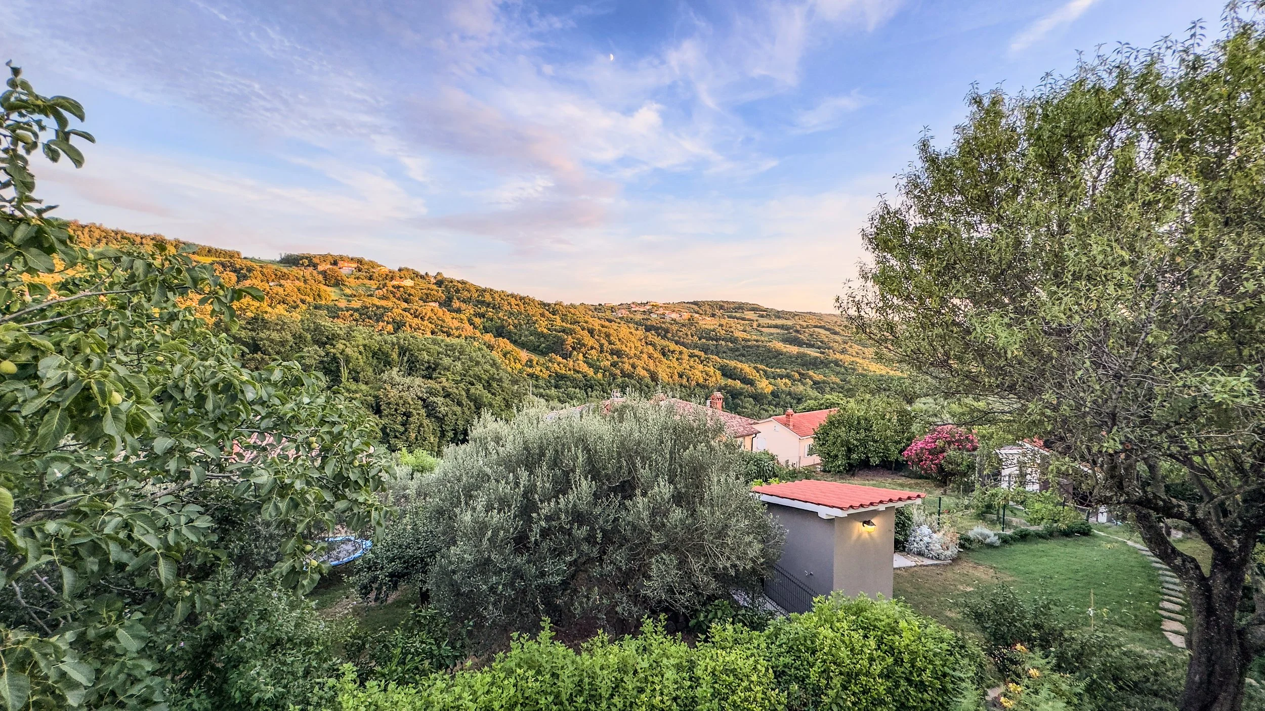 Scenic view of a hillside landscape with trees, houses with red roofs, lush greenery, and a sunset sky. Villa Domenico.