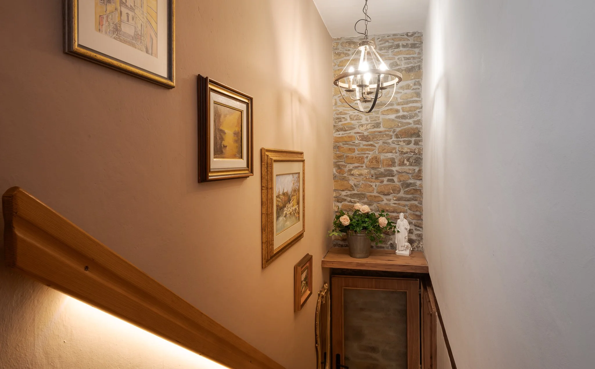 Interior view of a hallway with framed artwork on the left wall, a hanging chandelier, a brick accent wall at the end, a potted pink rose plant, and a white sculpture of a woman. The hallway has wooden flooring and a wooden door at the end.