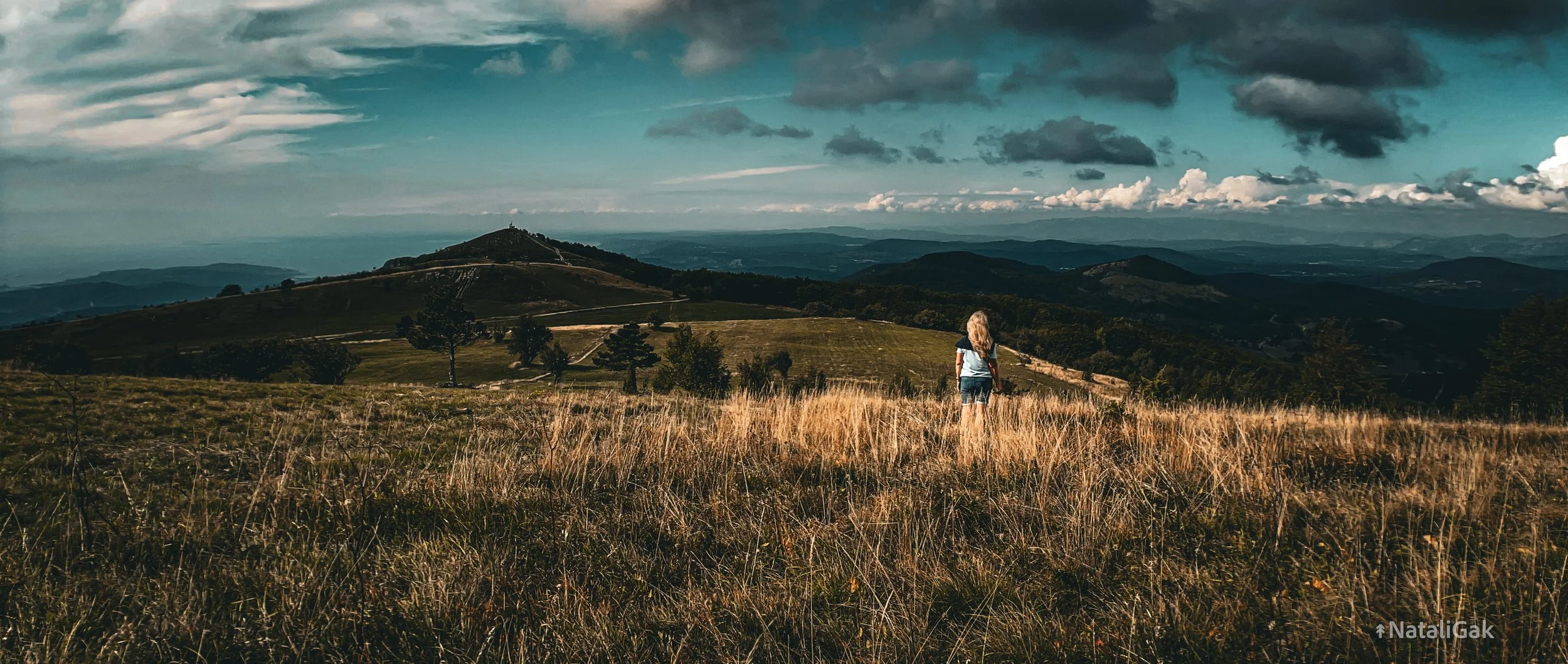 A woman walking through a grassy field under a cloudy sky with rolling hills in the distance.