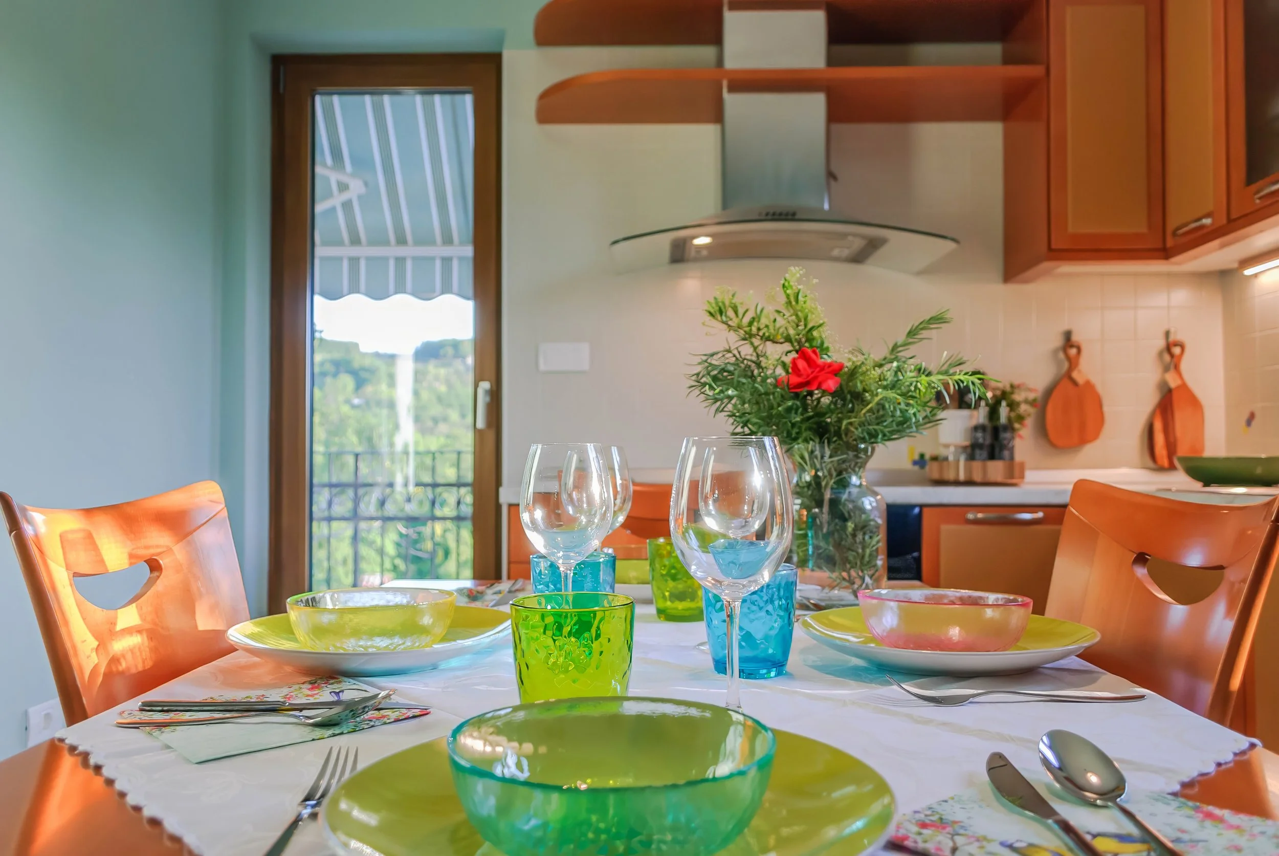 A dining table set with colorful glassware, bowls, and plates, with a centerpiece of flowers in a glass vase, in a bright kitchen with wooden chairs and a door leading outside. Located at Villa Domenico.