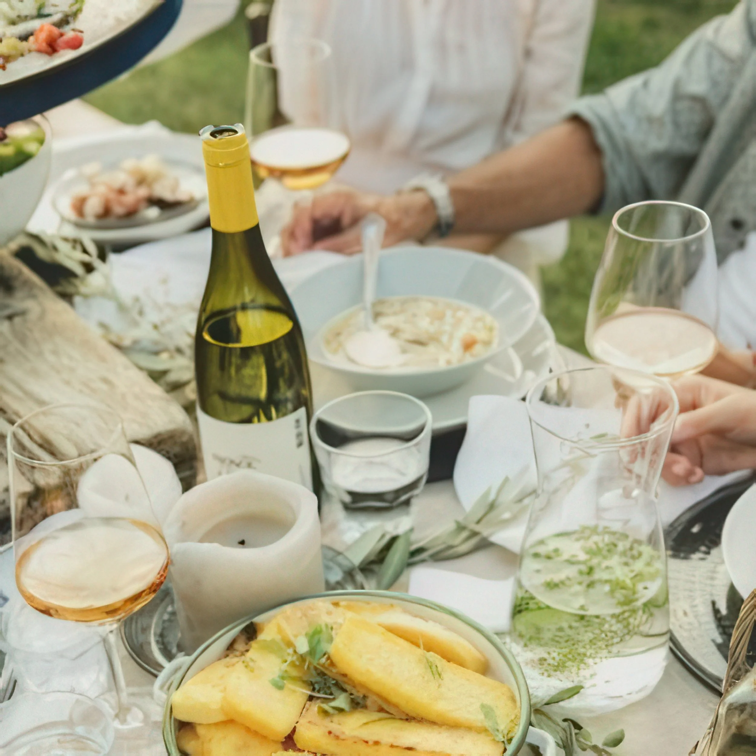 A white bowl filled with cooked shrimp garnished with herbs and lemon wedges, surrounded by other seafood dishes and beverages on a wooden table outdoors.