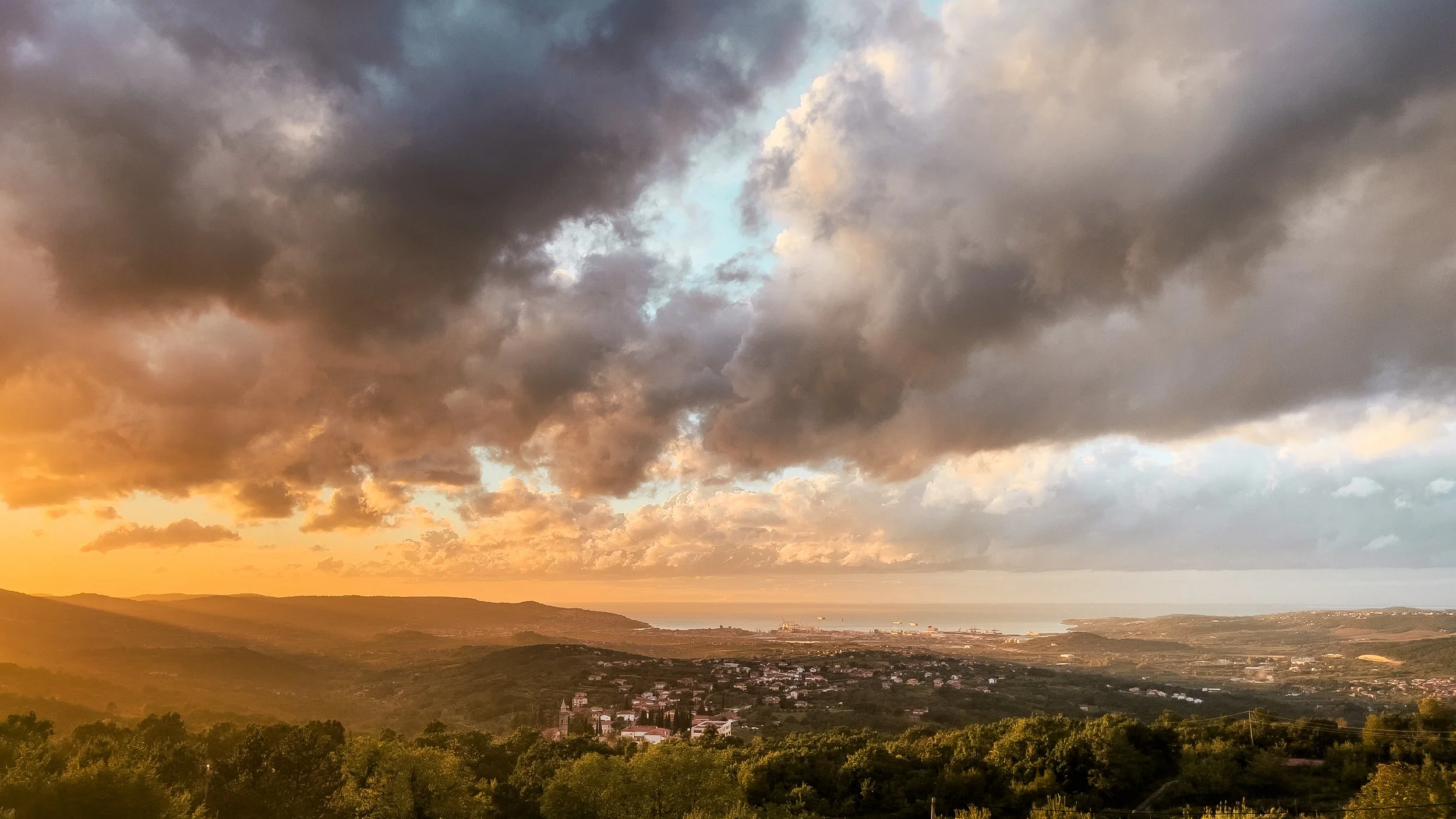 A scenic view of a sunset over a hilly landscape with dark clouds in the sky and a town in the distance near the coast.