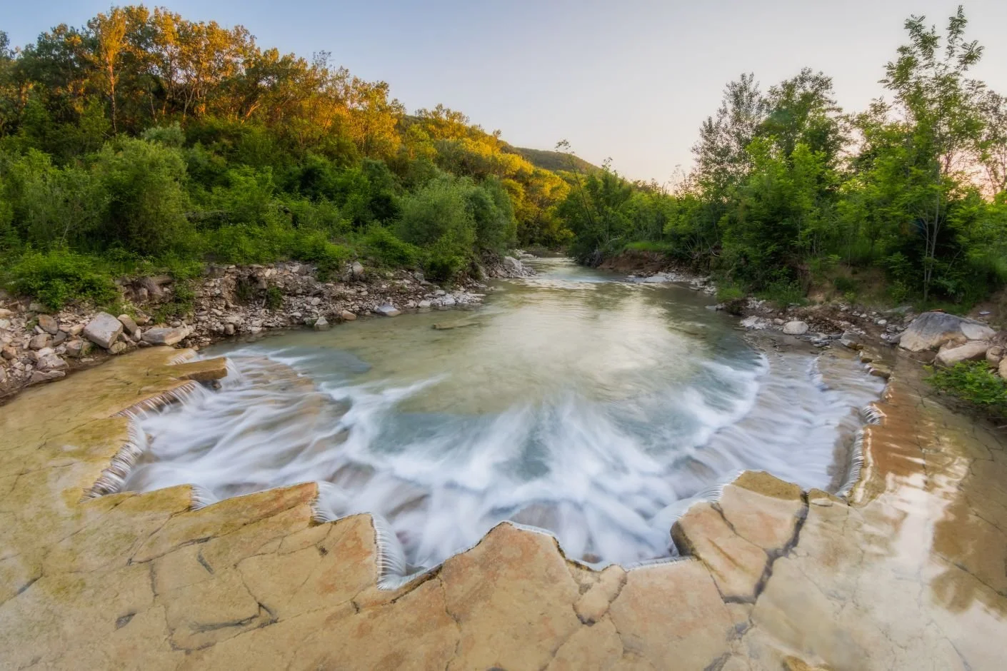 A natural hot spring with water bubbling over rocks, surrounded by green trees on a hillside, during sunset.