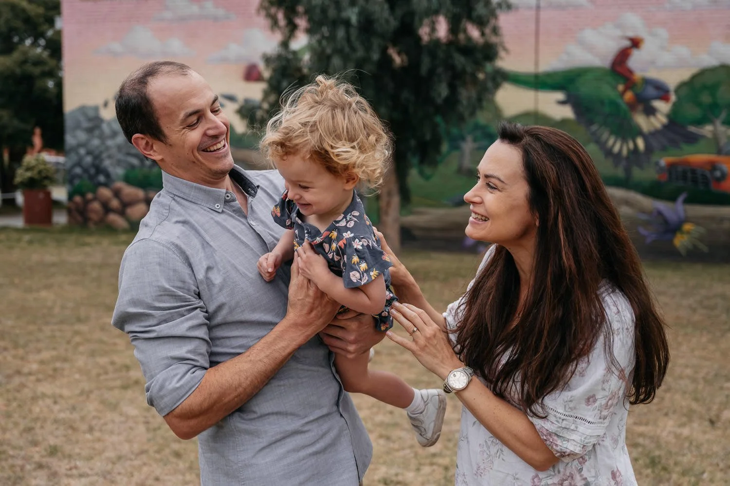A happy family outdoors with a man, woman, and a young child. The man and woman are smiling at the child, who is being held up and is laughing. The background features trees and a colorful mural with a bird and a person riding a bird.