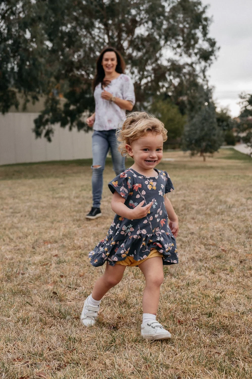 A young girl with curly blonde hair, wearing a navy floral dress and white sneakers, is running on a grassy field. In the background, a woman with long dark hair, wearing a white top and ripped jeans, is smiling and walking toward her, with trees and