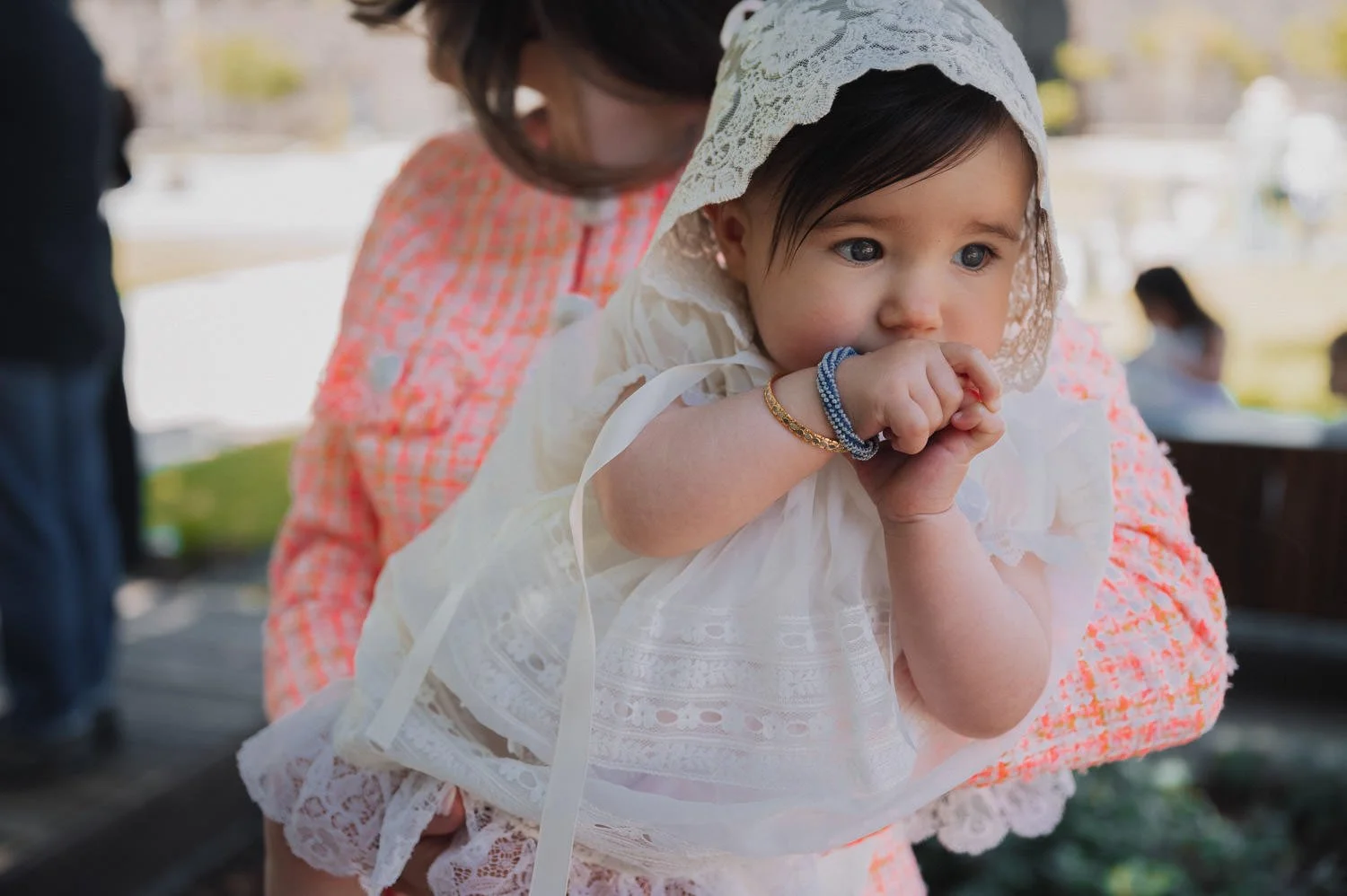 A young girl dressed in a white lace dress and bonnet, with blue eyes and dark hair, is being held by an adult. The girl is sucking on her fingers and wearing bracelets on her wrists.
