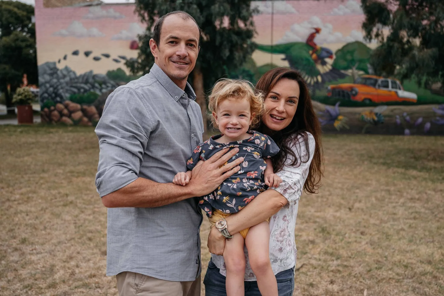 A family of three consisting of a man, woman, and young girl smiling and standing outdoors in front of a colorful mural.