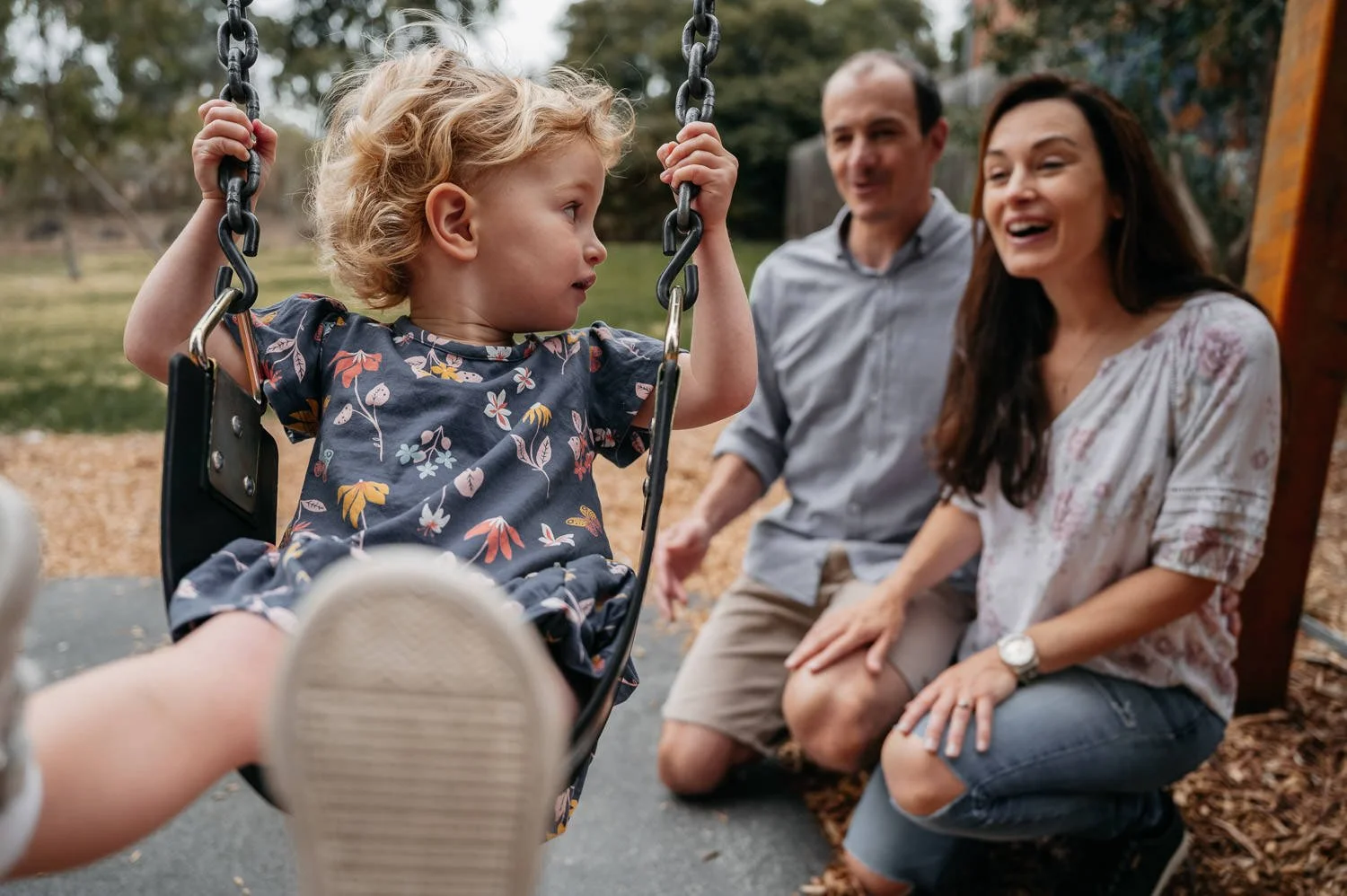 A young girl on a swing talking to a man and woman kneeling on the ground, in a park setting.